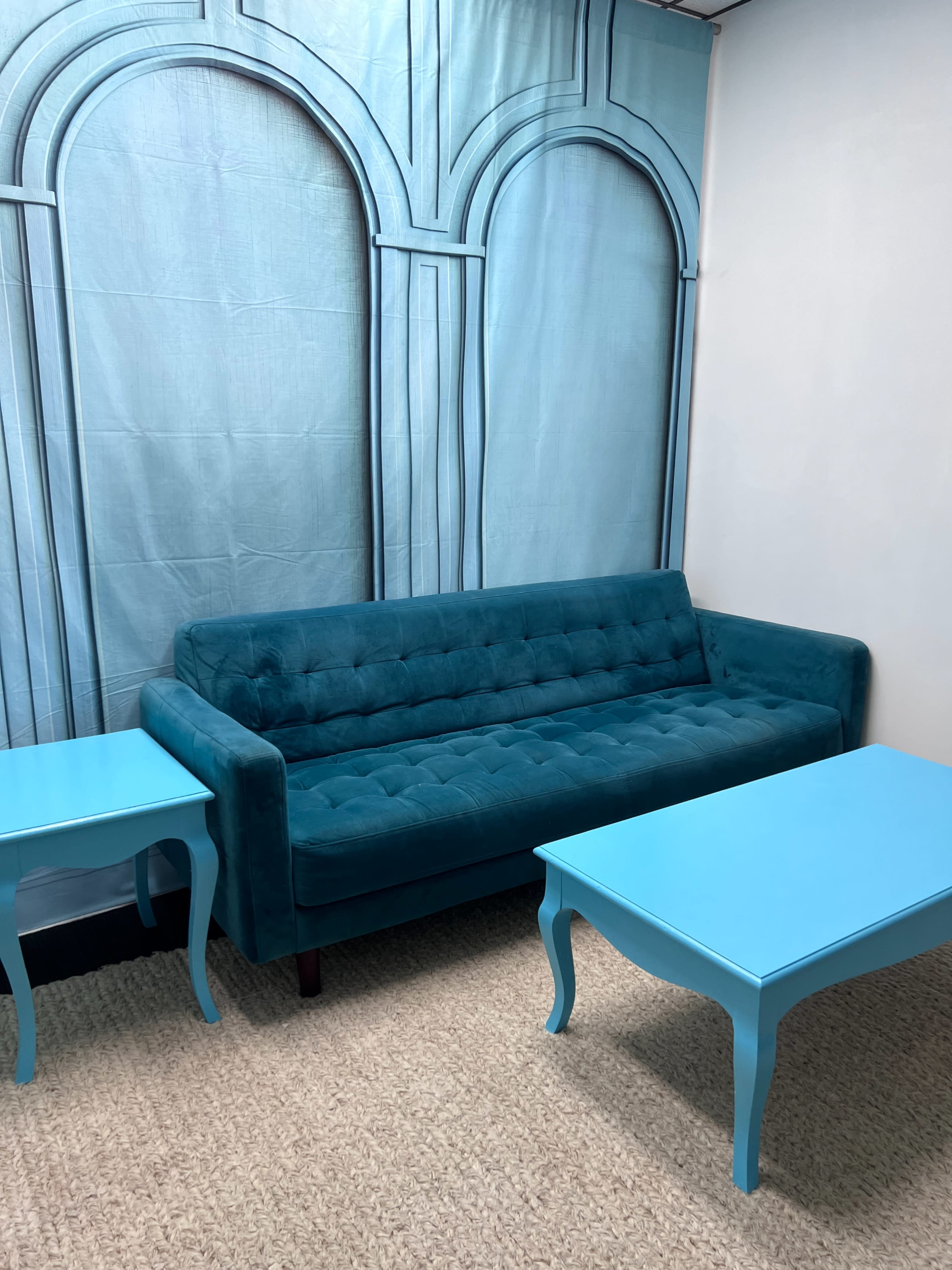 The image shows a teal couch placed against a matching blue backdrop, with two blue side tables on either side.