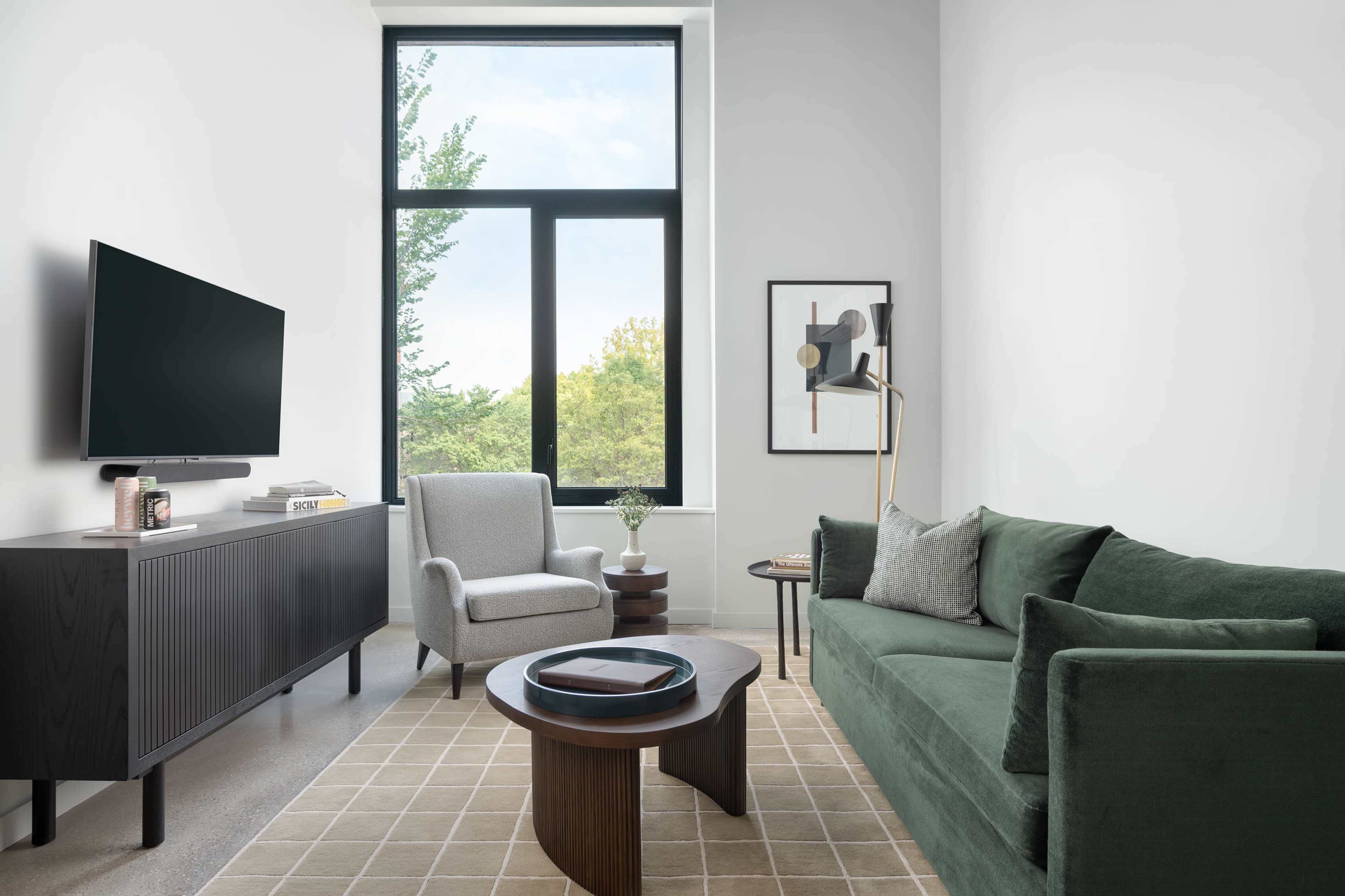 The image features a modern living room with a green couch, an armchair, a coffee table, a television, and a large window providing a view of greenery outside.