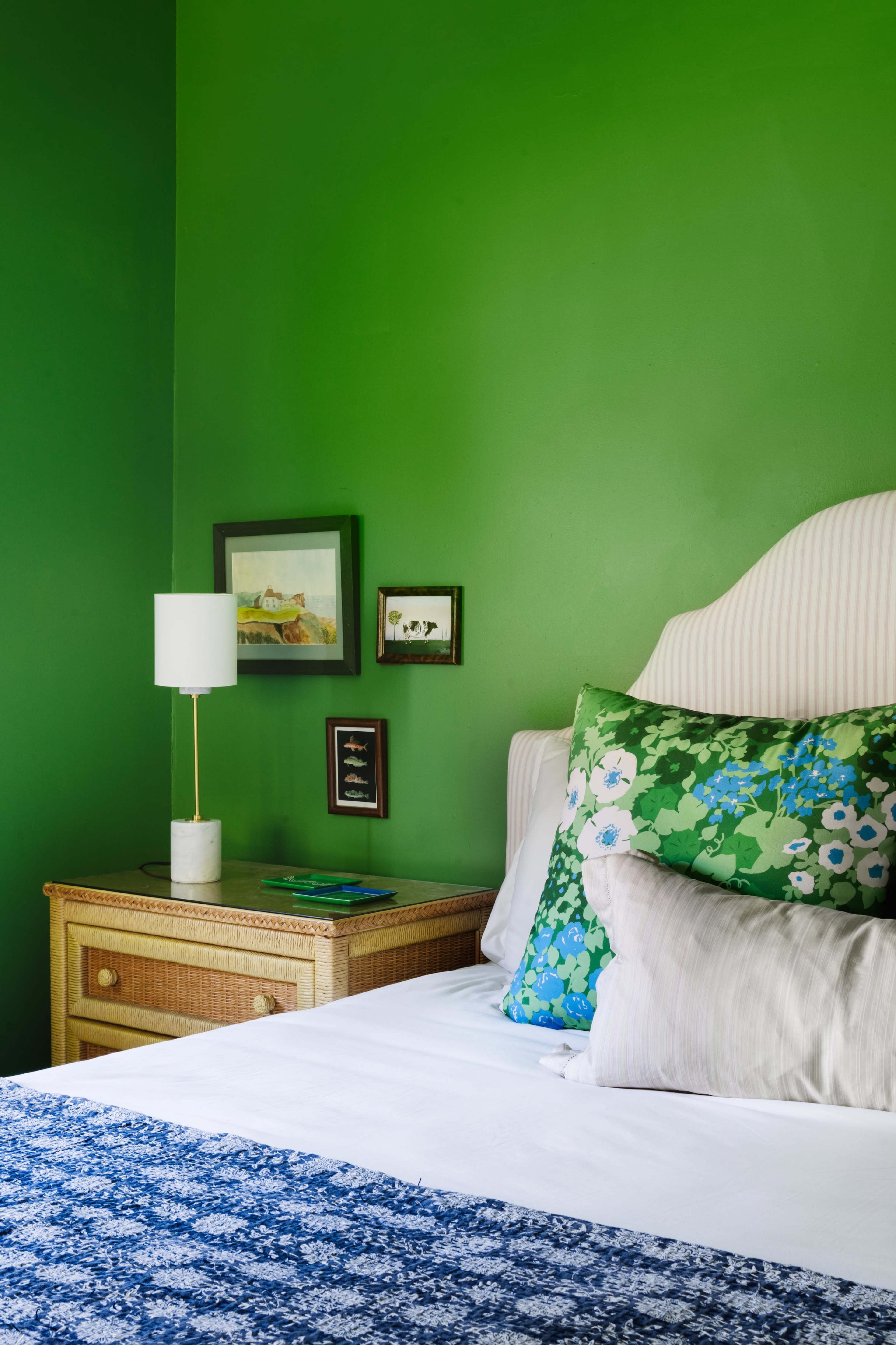 A bedroom with a green accent wall, a bed adorned with floral pillows, and a bedside table holding a lamp and framed pictures.