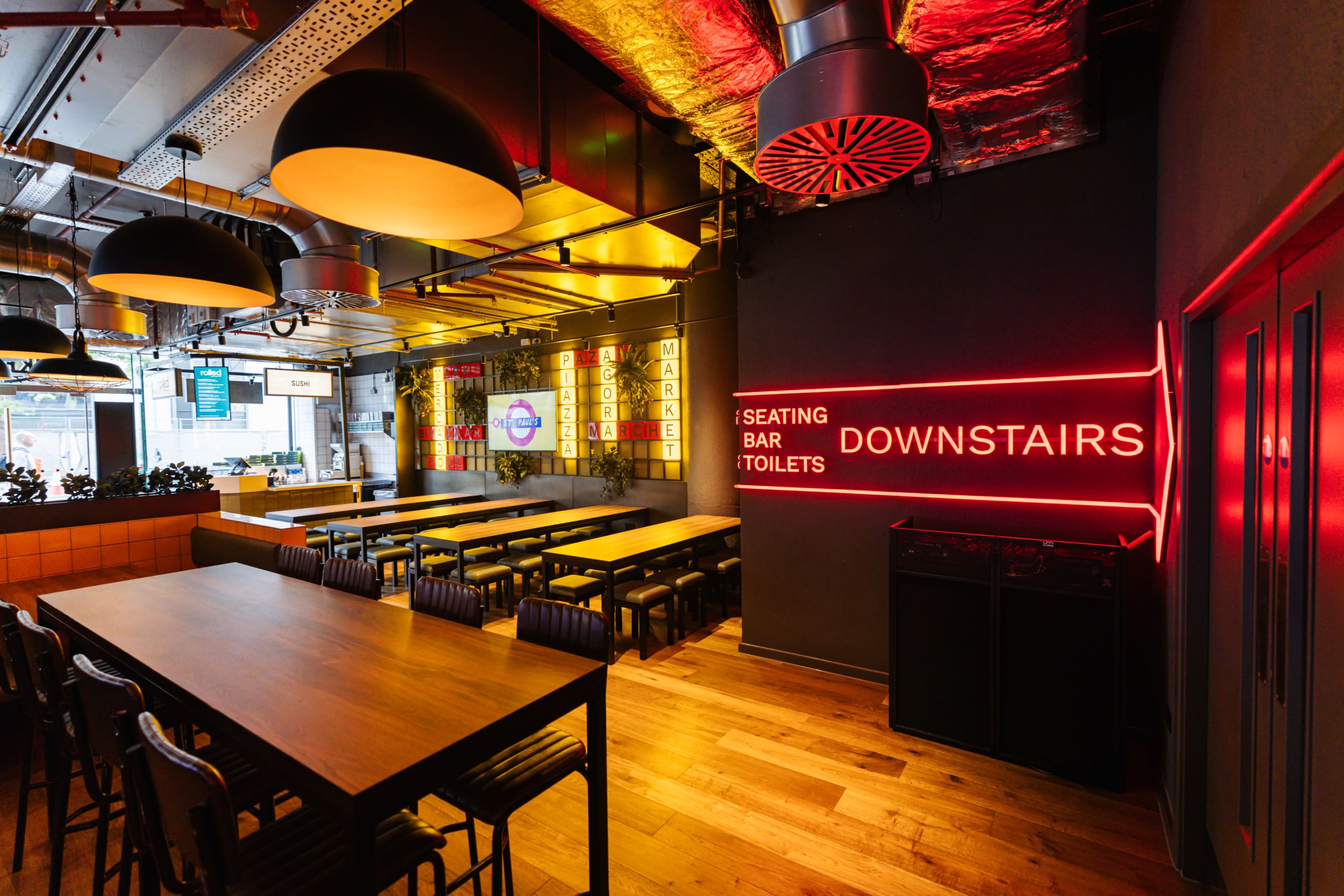 The image shows a modern dining area with long tables, bar seating, and illuminated signs directing to the downstairs area and facilities like the bar and toilets.