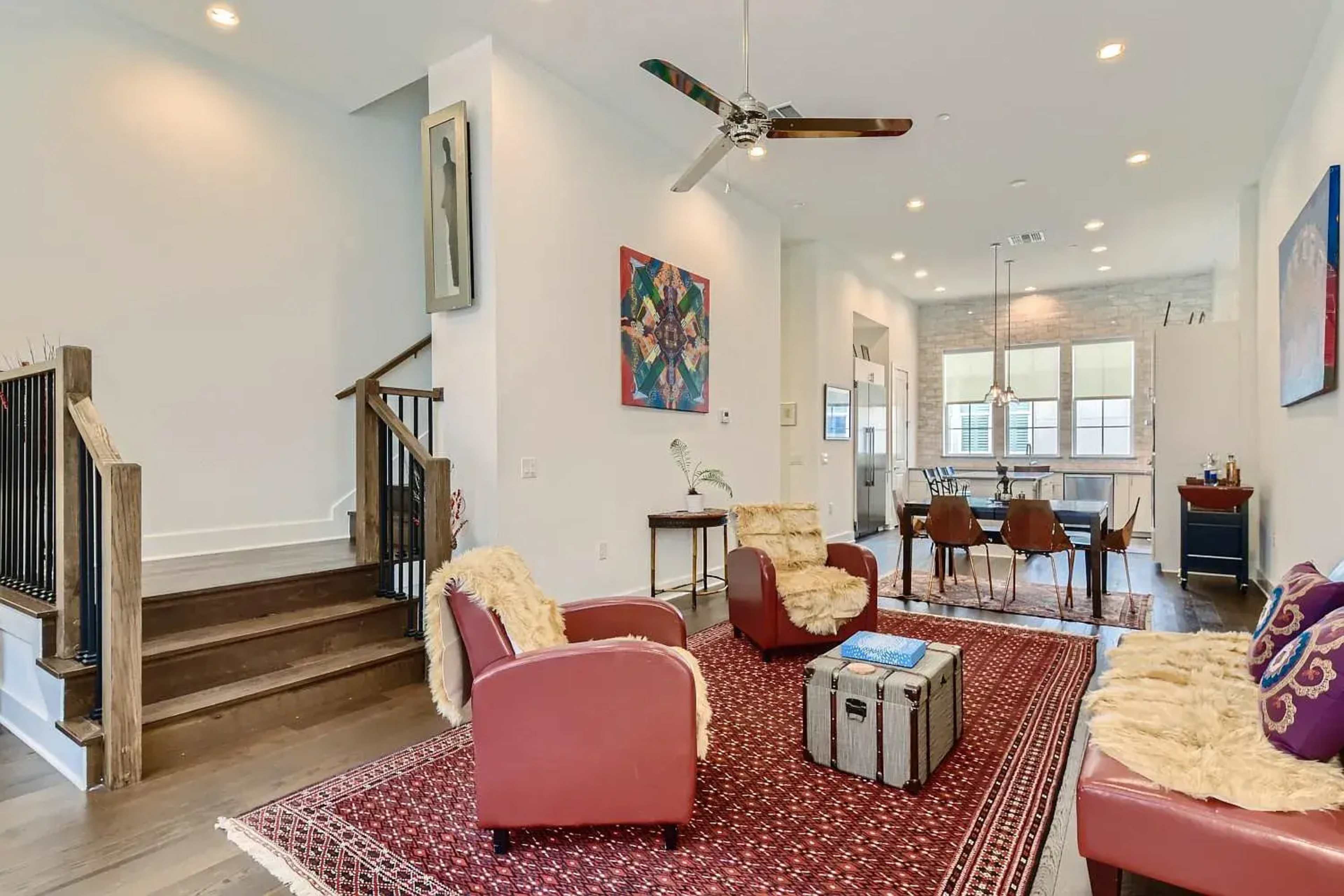 The image shows a living room with hardwood floors, a red patterned rug, two red armchairs, a coffee table, and a staircase leading to an upper level.