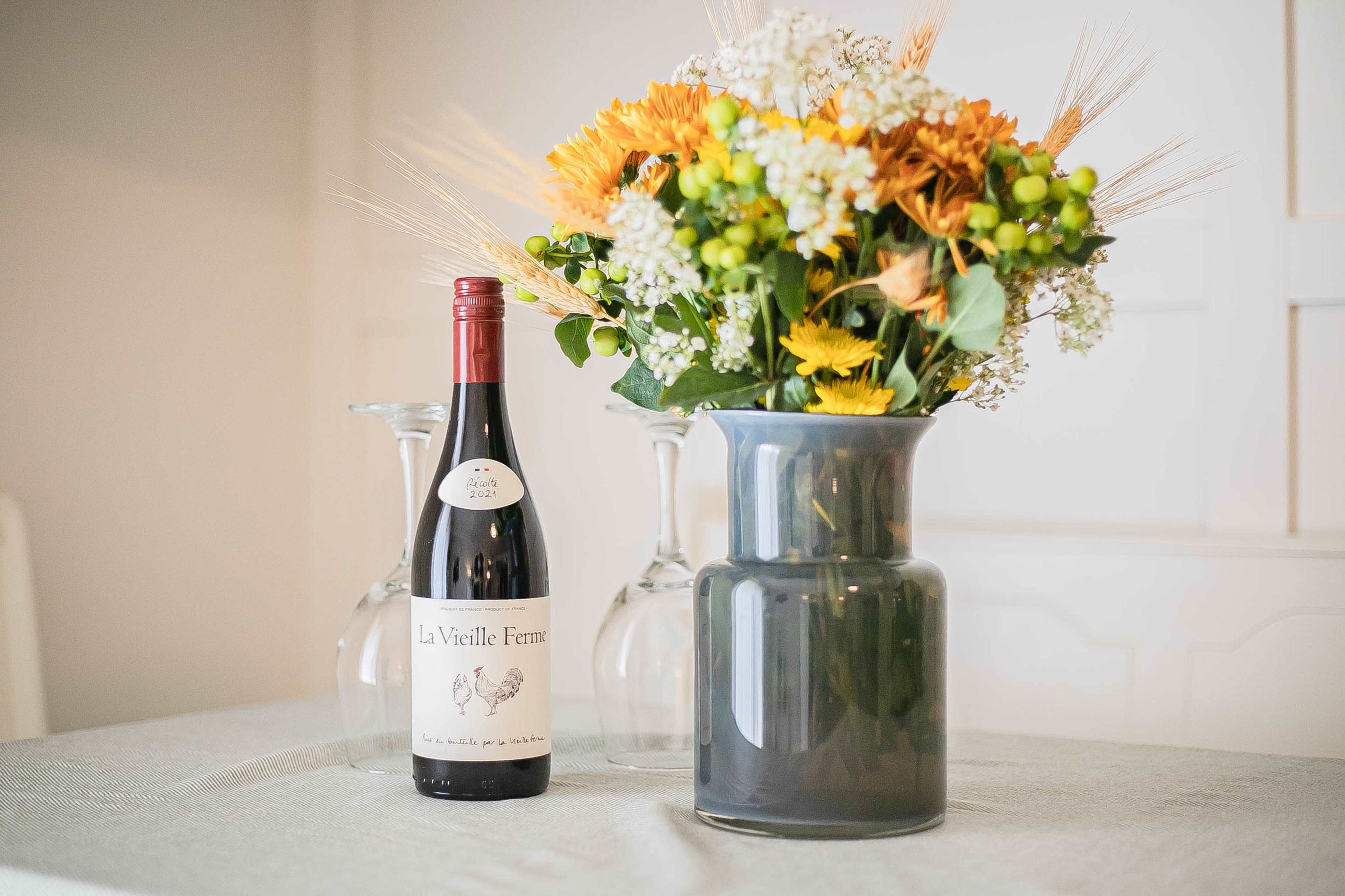 A bottle of red wine stands beside a bouquet of colorful flowers in a glass vase on a table.