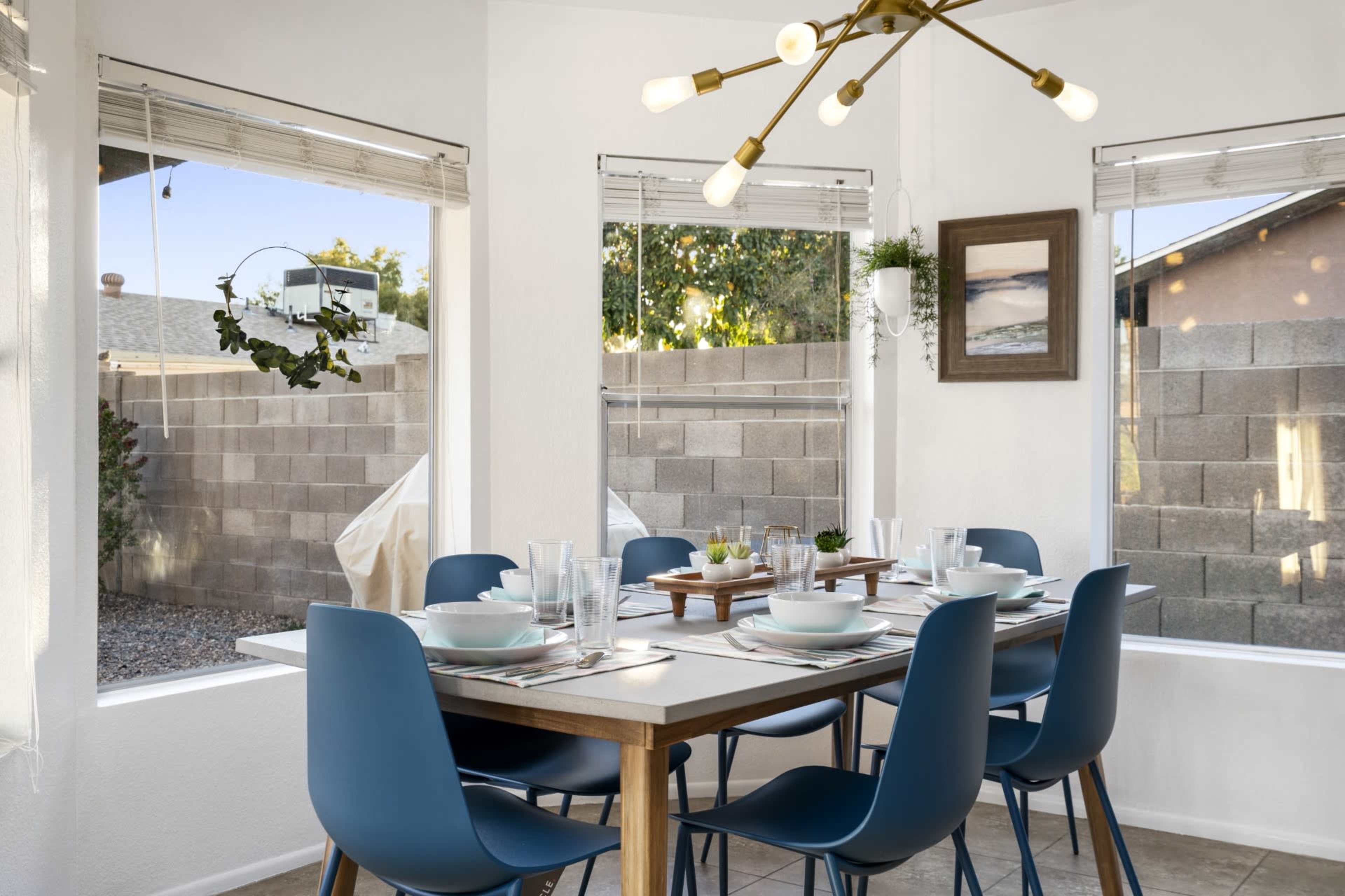 A modern dining area features a wooden table set for eight with blue chairs, surrounded by large windows showcasing a landscaped backyard.