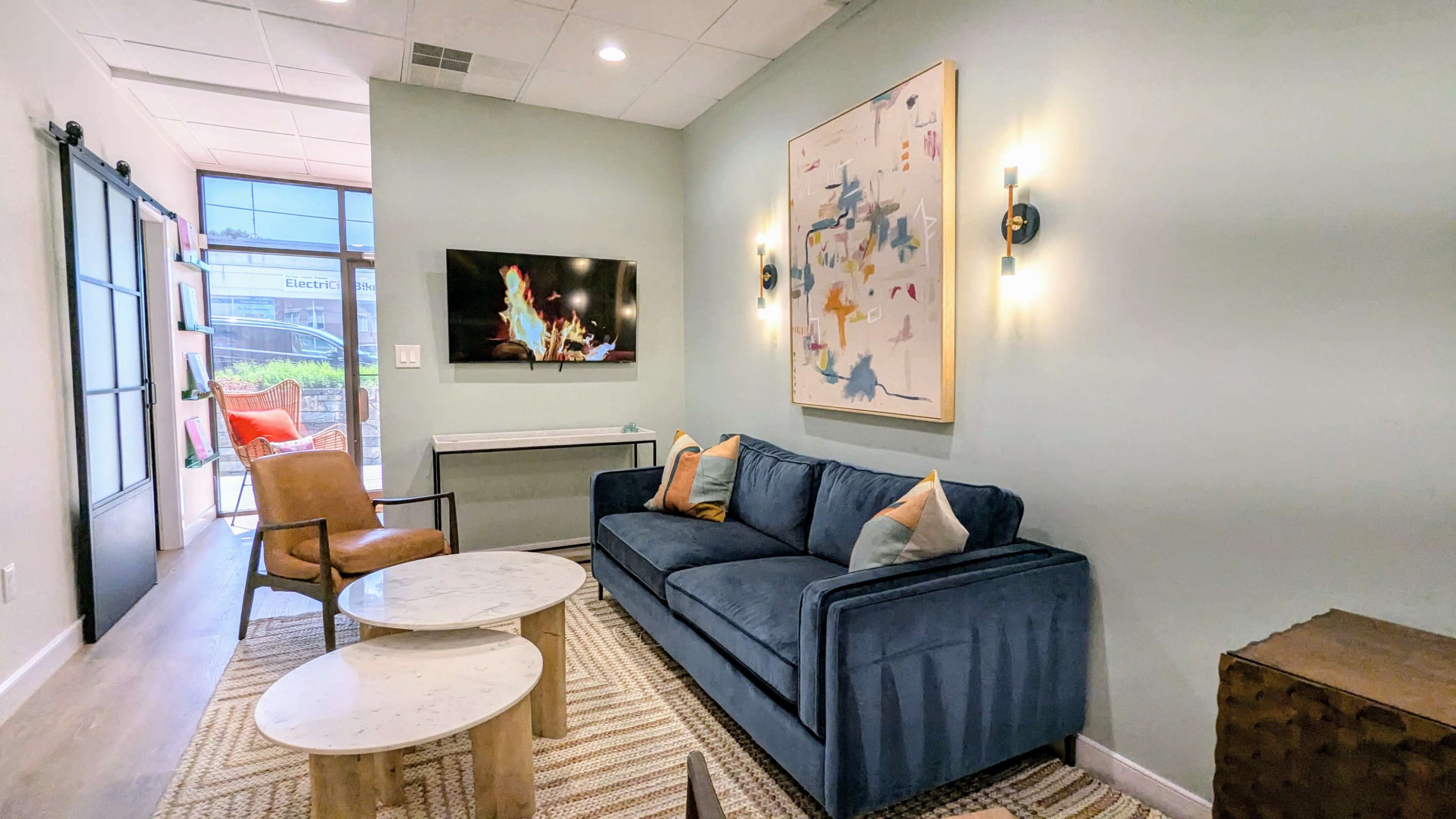 A modern sitting area with a blue couch, two coffee tables, a wall-mounted television, and a piece of abstract art on the wall.