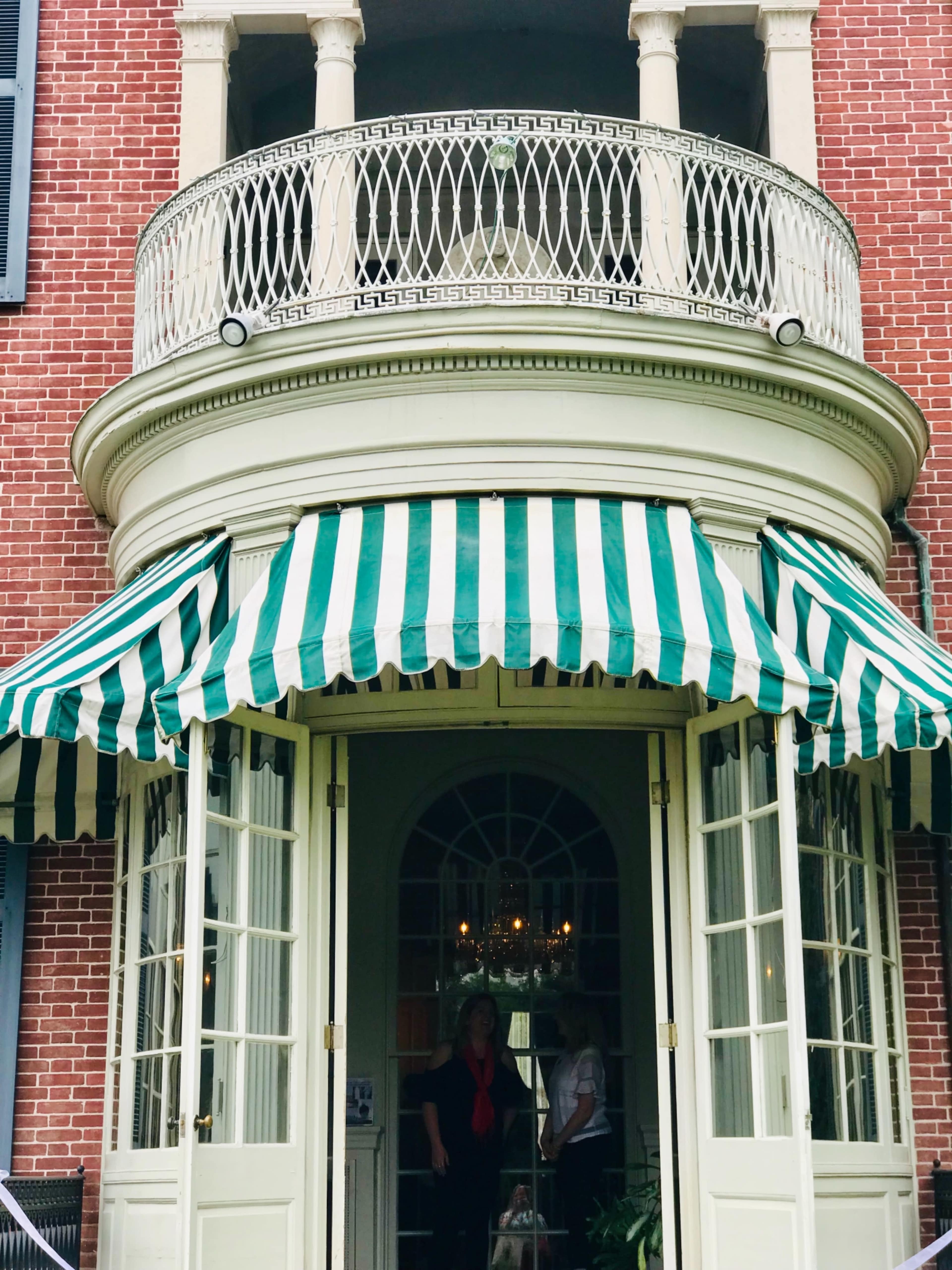 A woman stands under a green and white striped awning at the entrance of a brick building with a rounded balcony.