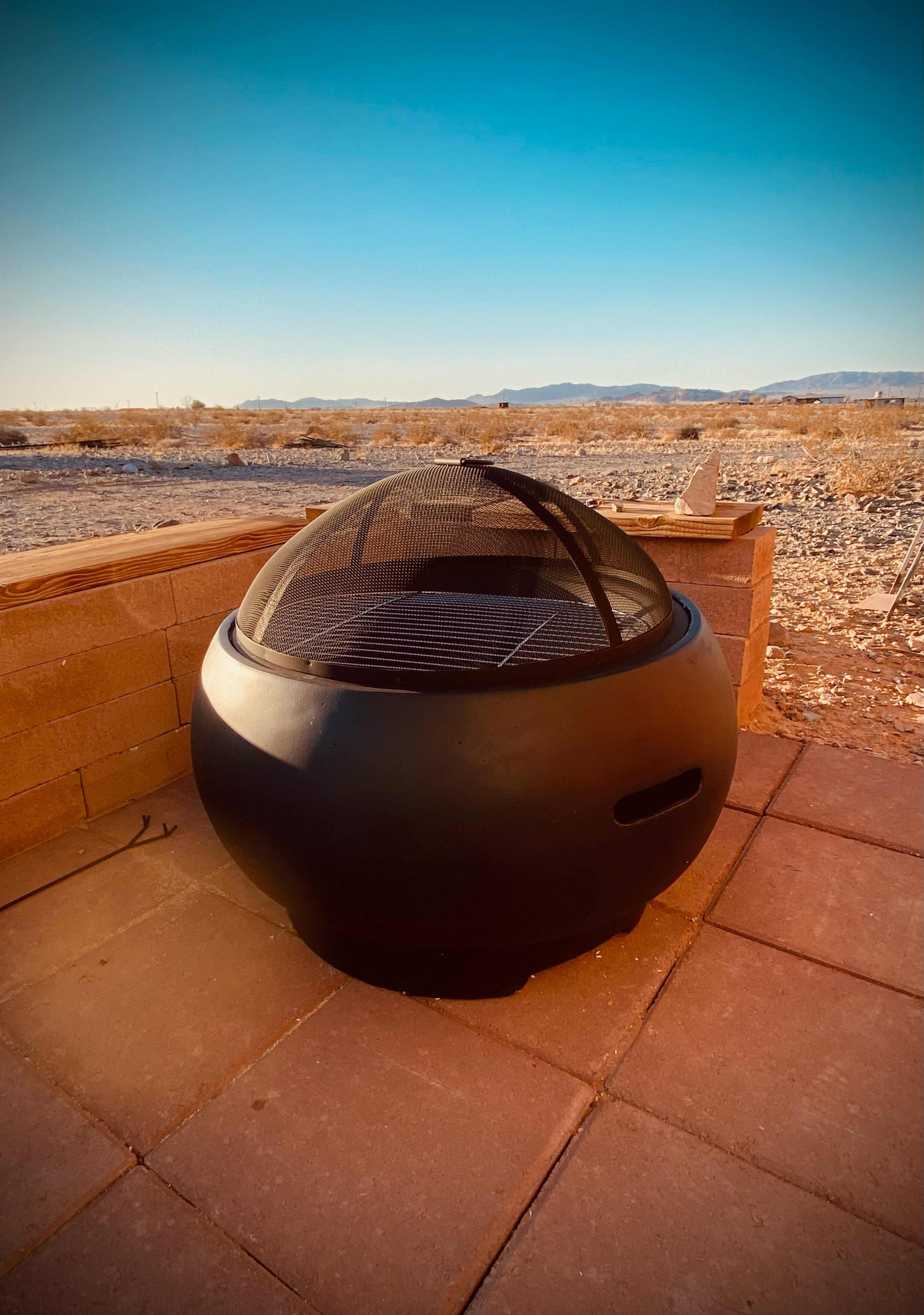 A black, spherical fire pit with a mesh lid sits on a stone patio against a desert landscape.