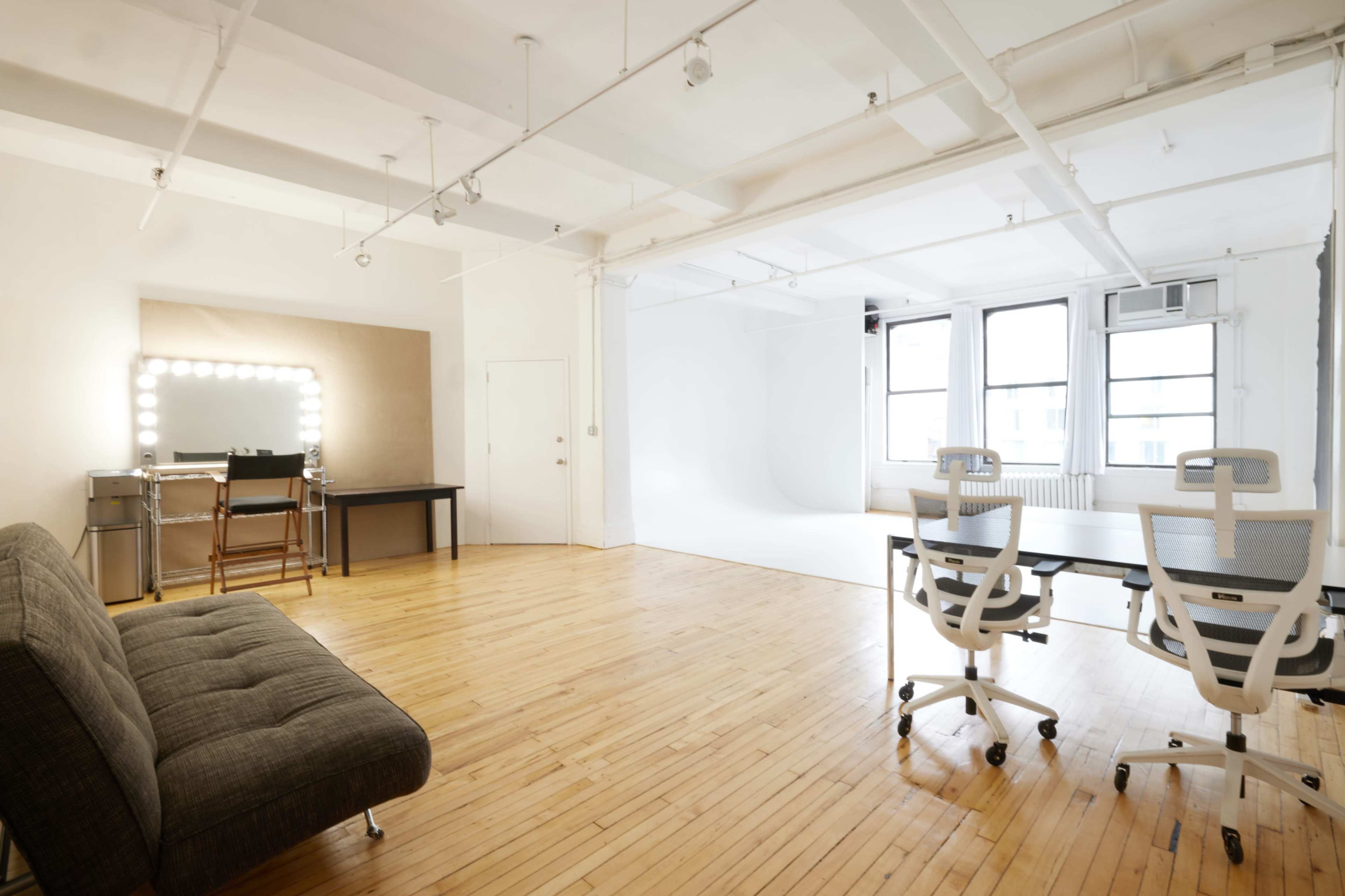 A spacious, well-lit studio featuring a seating area with a couch, a dressing table with lights, and two office chairs positioned at a table.