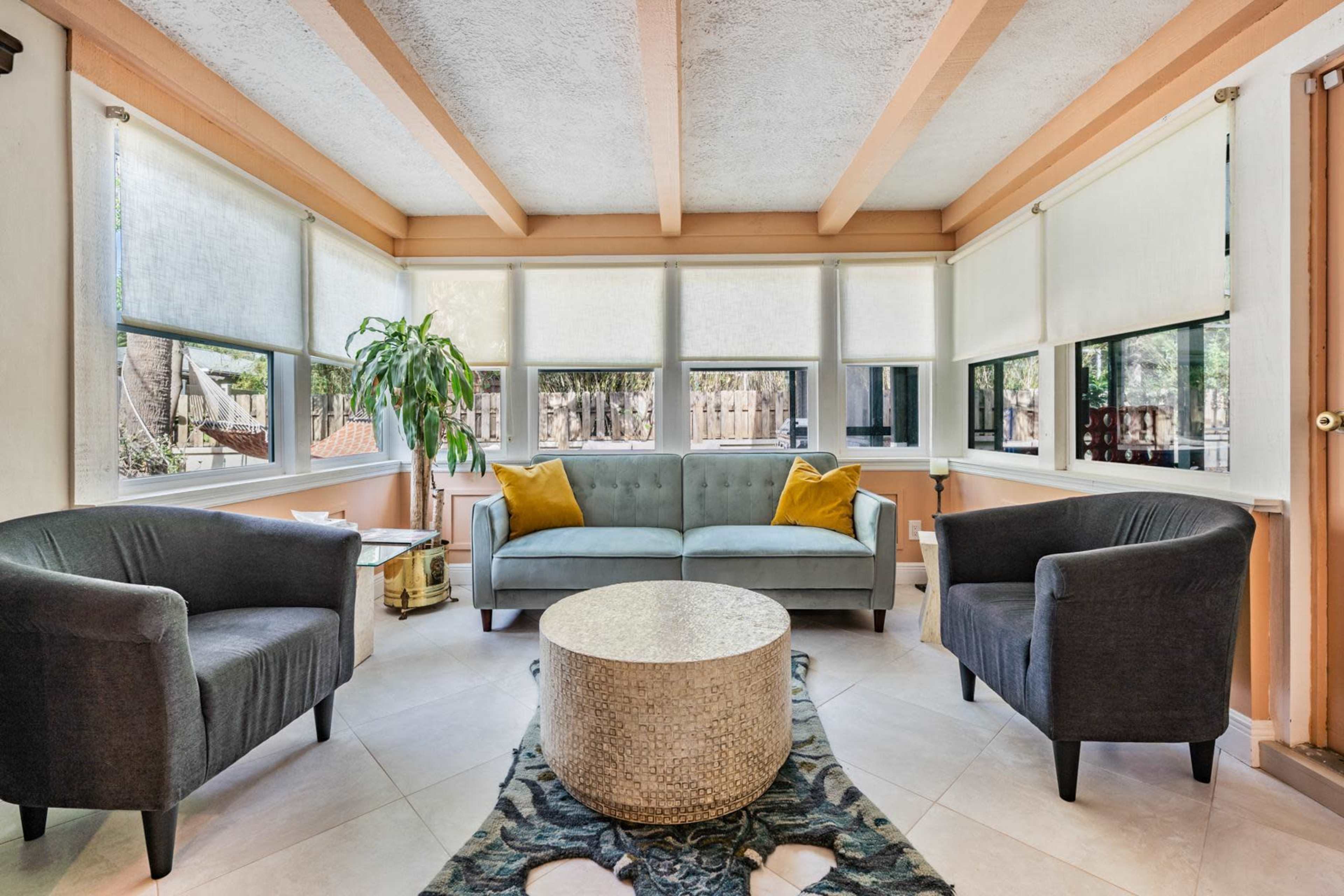 A sunlit sitting area features a gray sofa with yellow cushions, two matching armchairs, and a round stone coffee table, surrounded by large windows.