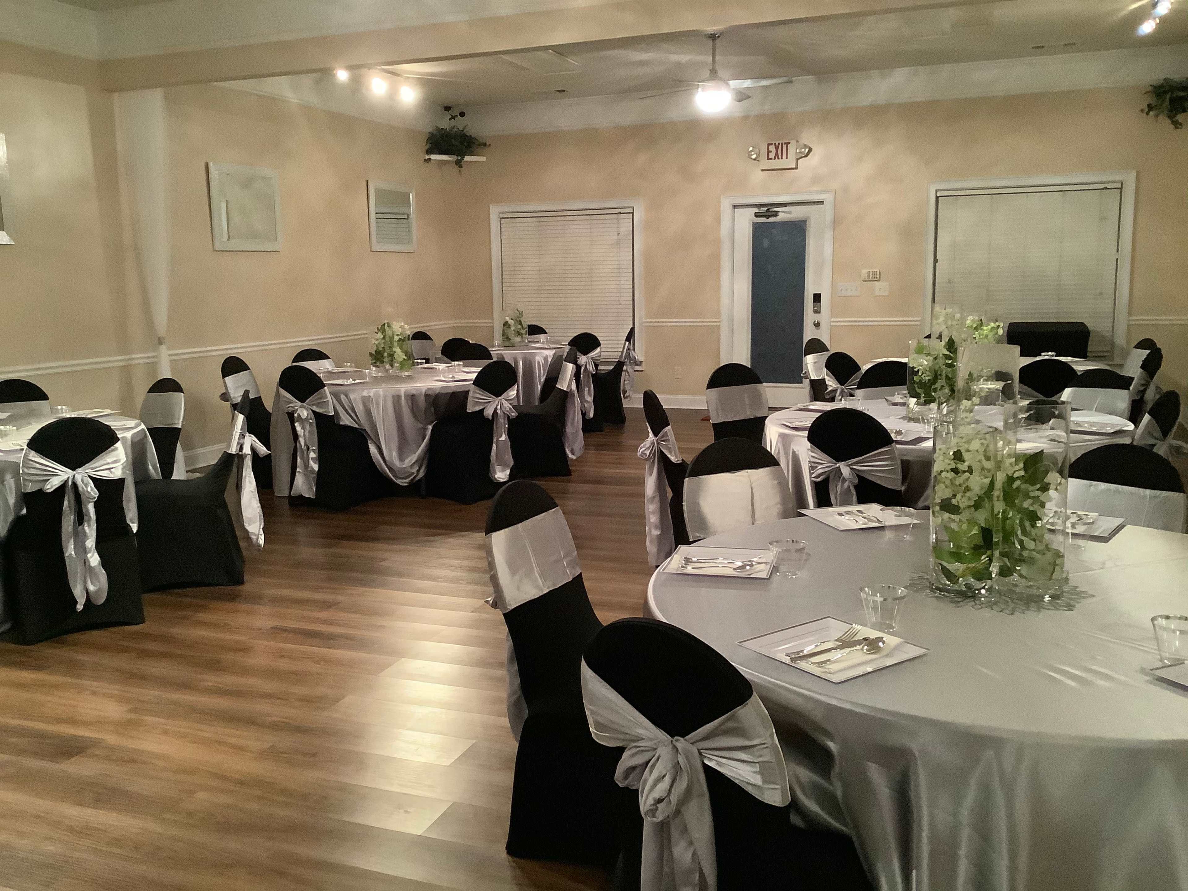 A decorated event space with round tables covered in silver and black linens, each set with white chairs adorned with silver bows.