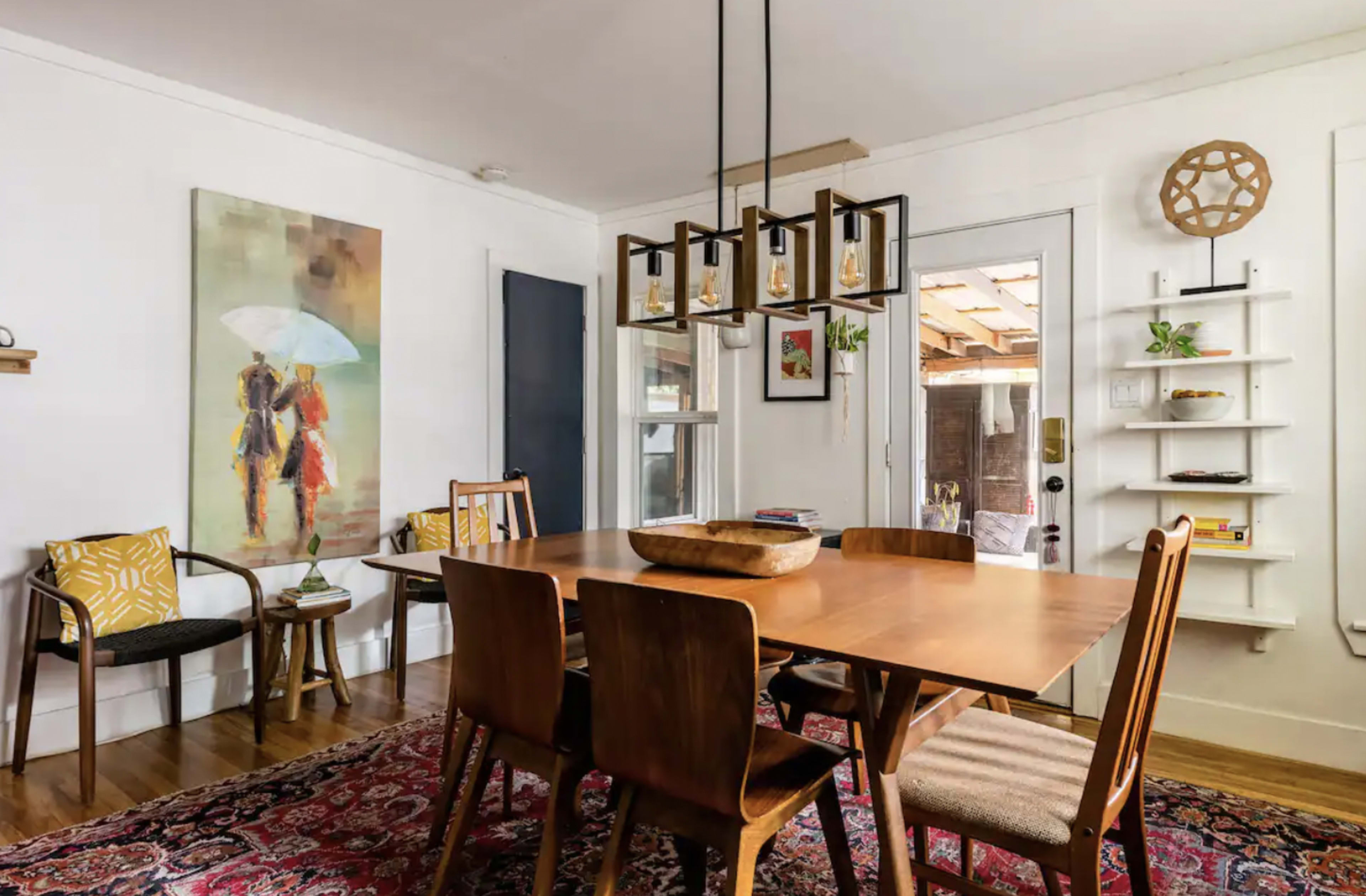 A wooden dining table surrounded by chairs is positioned in a room with a colorful painting on the wall and a doorway leading to a patio.