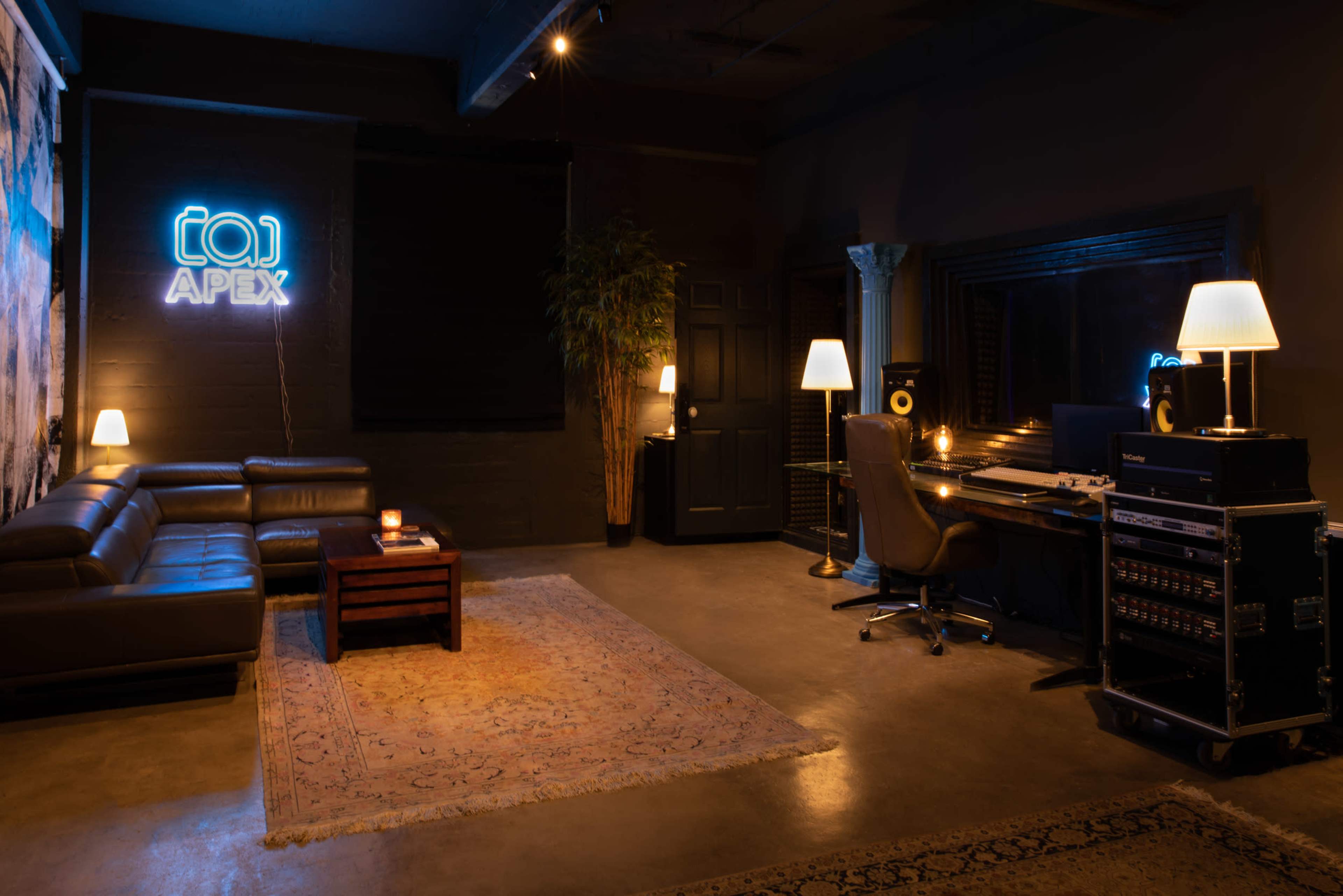 A dimly lit music studio features a leather sofa, a wooden table with a candle, a desk with recording equipment, and a neon "APEX" sign on the wall.