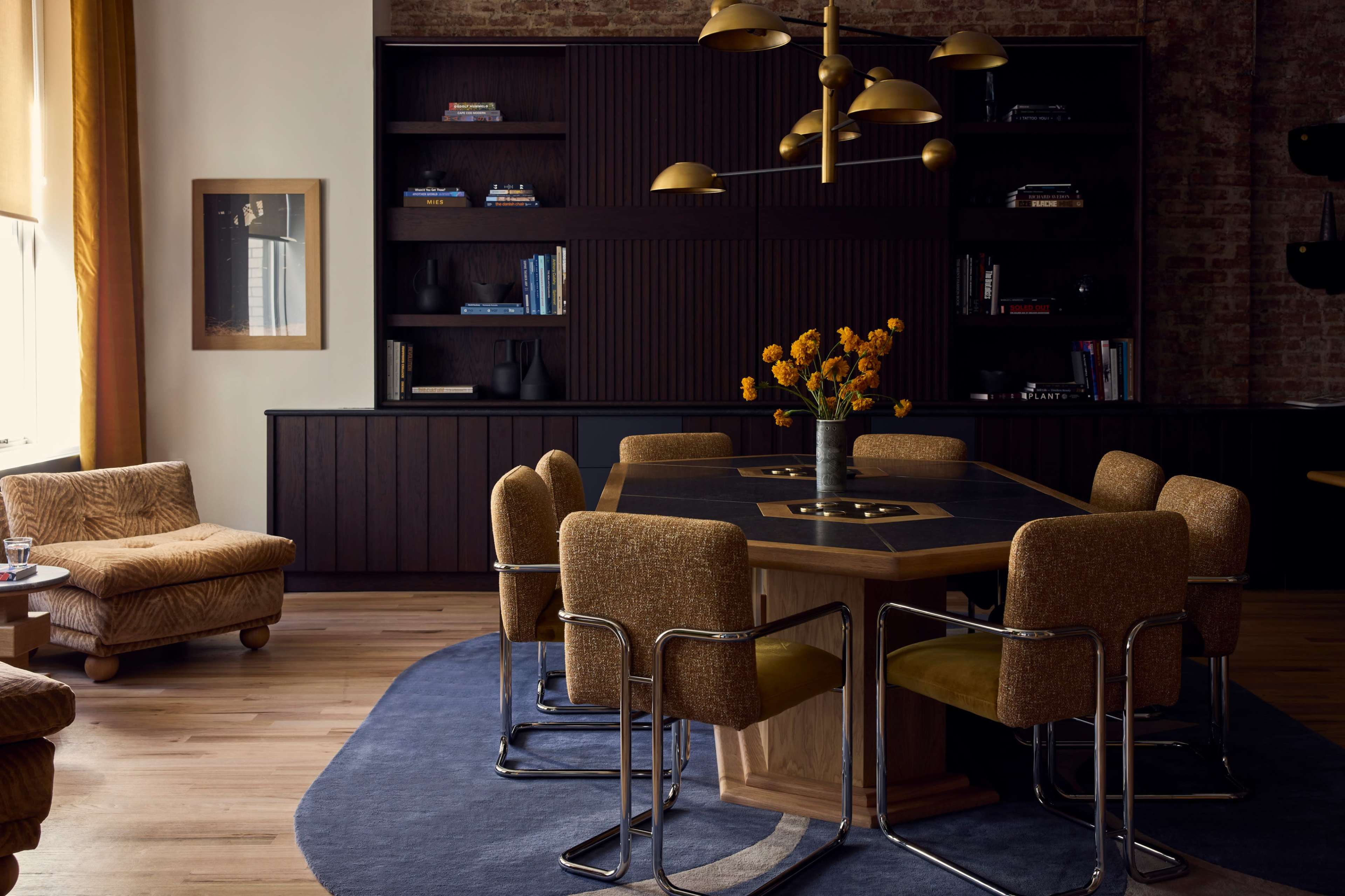 A modern dining area features a wooden table surrounded by upholstered chairs, with a vase of flowers as the centerpiece and a bookshelf lined with books in the background.