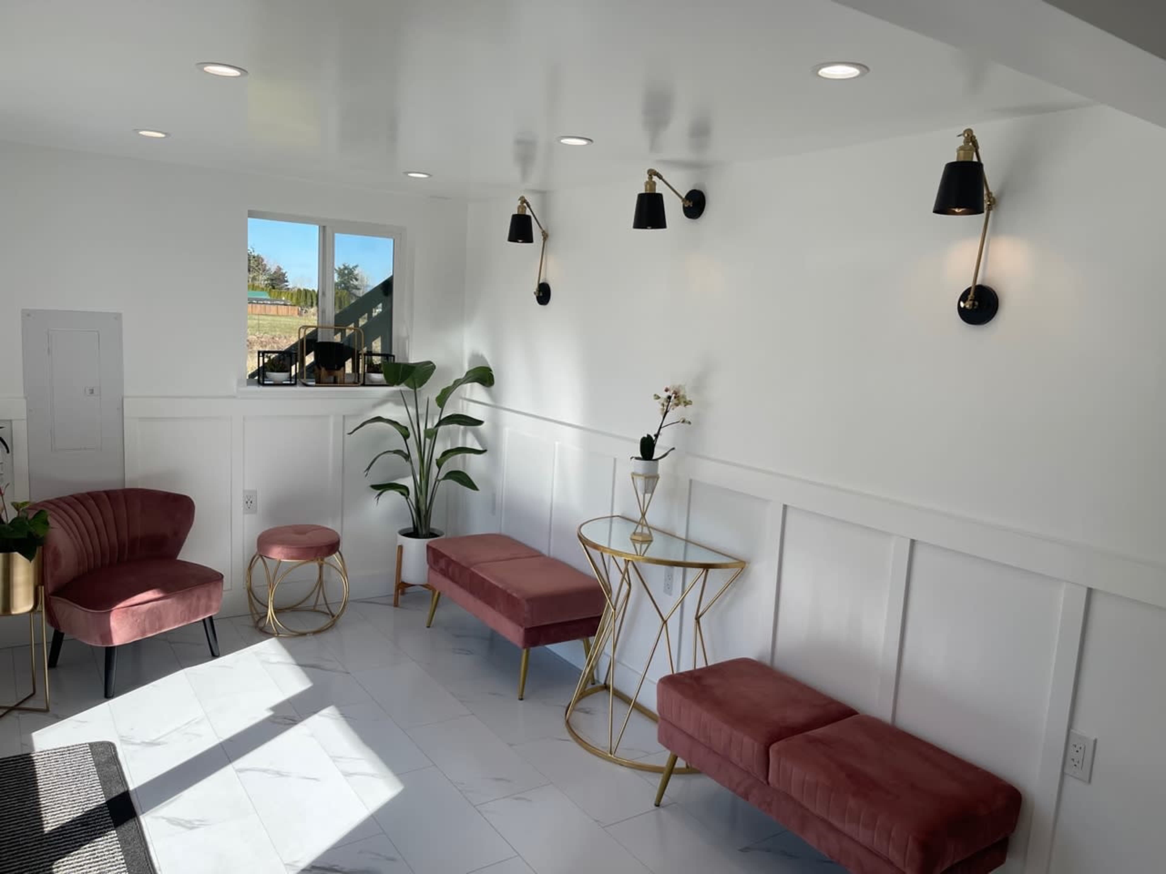 A bright room with white walls and a tiled floor, furnished with several pink seating options, a gold side table, and wall-mounted black lamps.