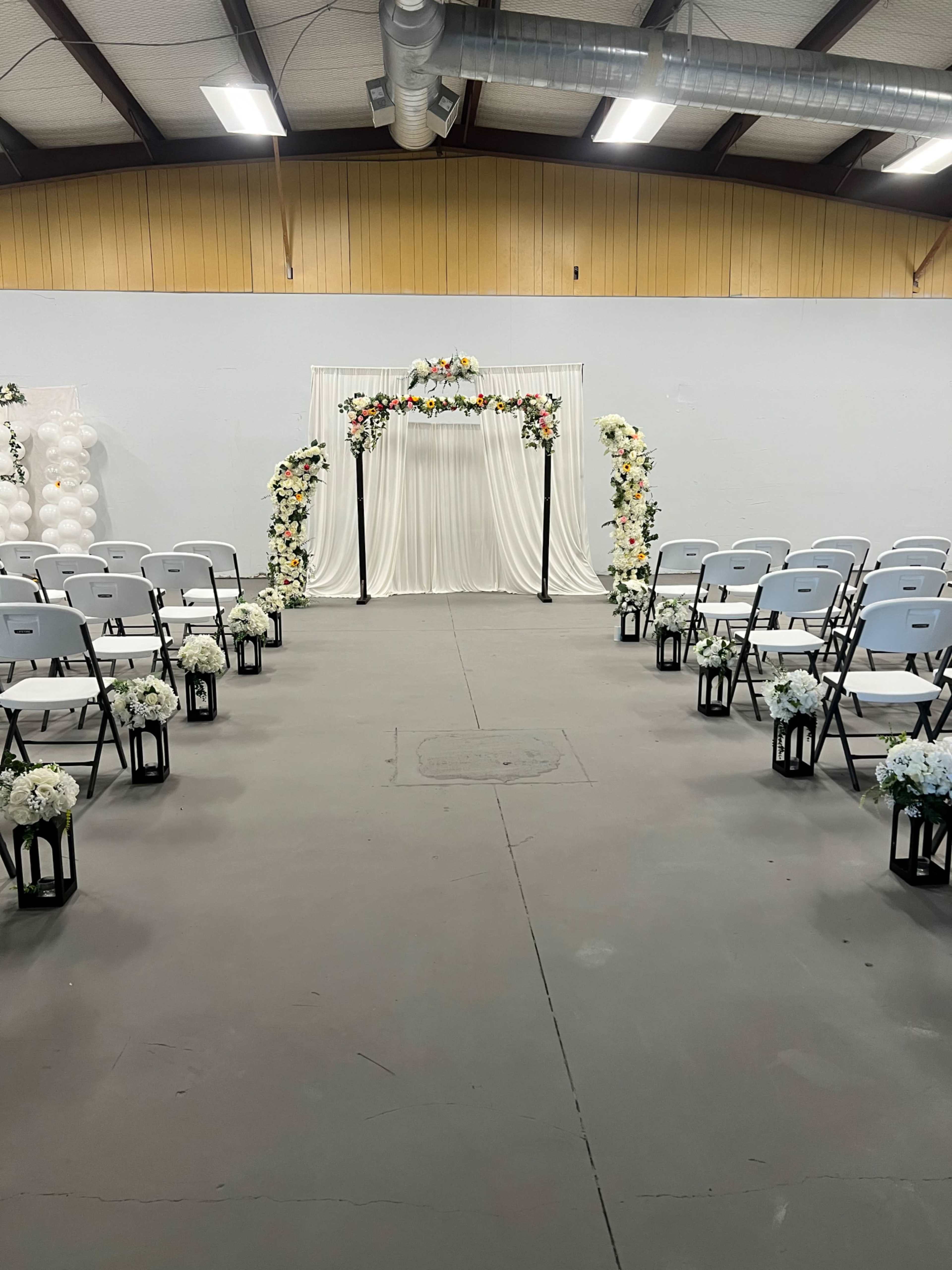A wedding ceremony setup features rows of empty chairs facing a floral arch draped in white fabric at the front.