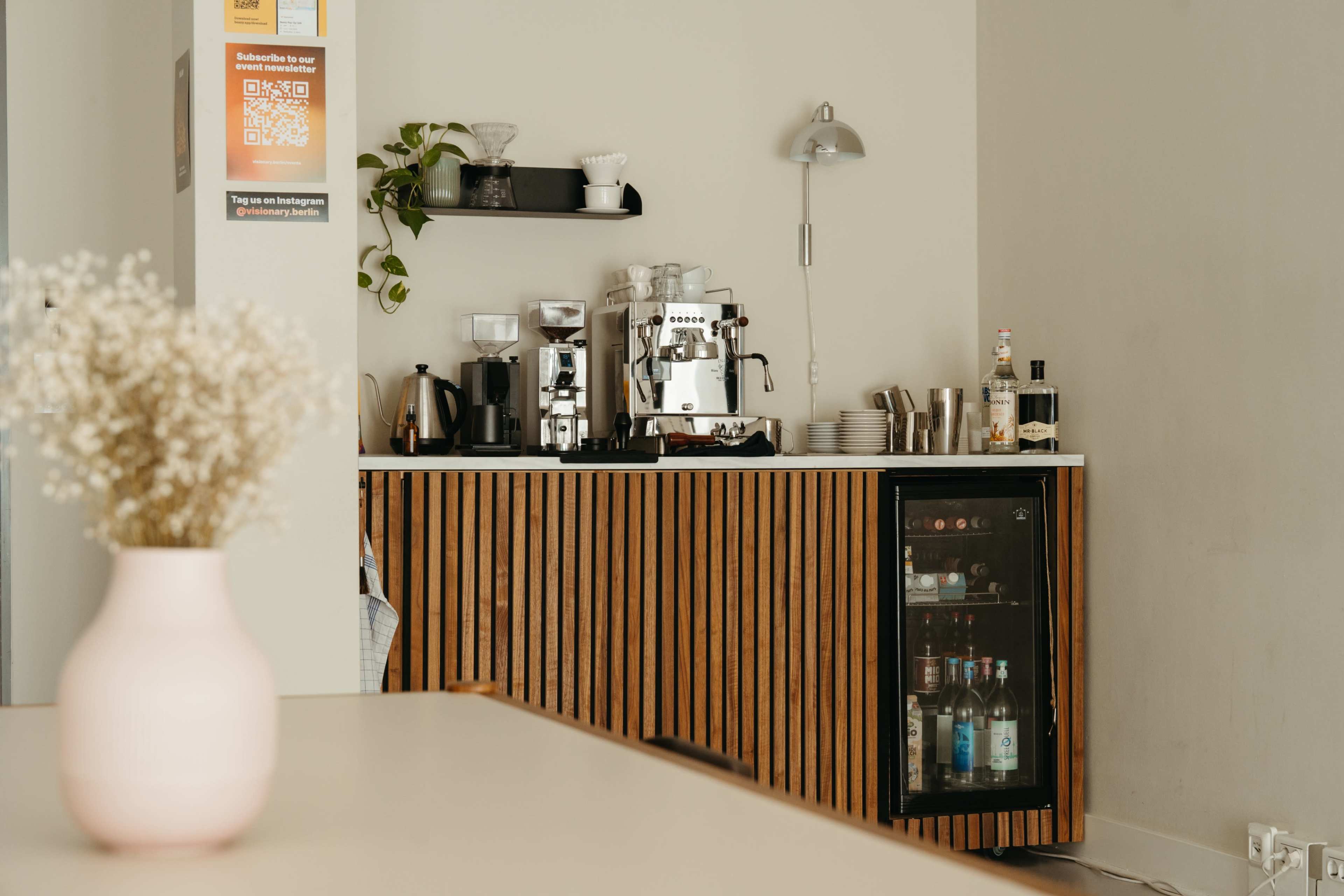 A wooden countertop with a coffee machine, various utensils, and a glass-fronted cabinet displaying bottles is set against a plain wall.