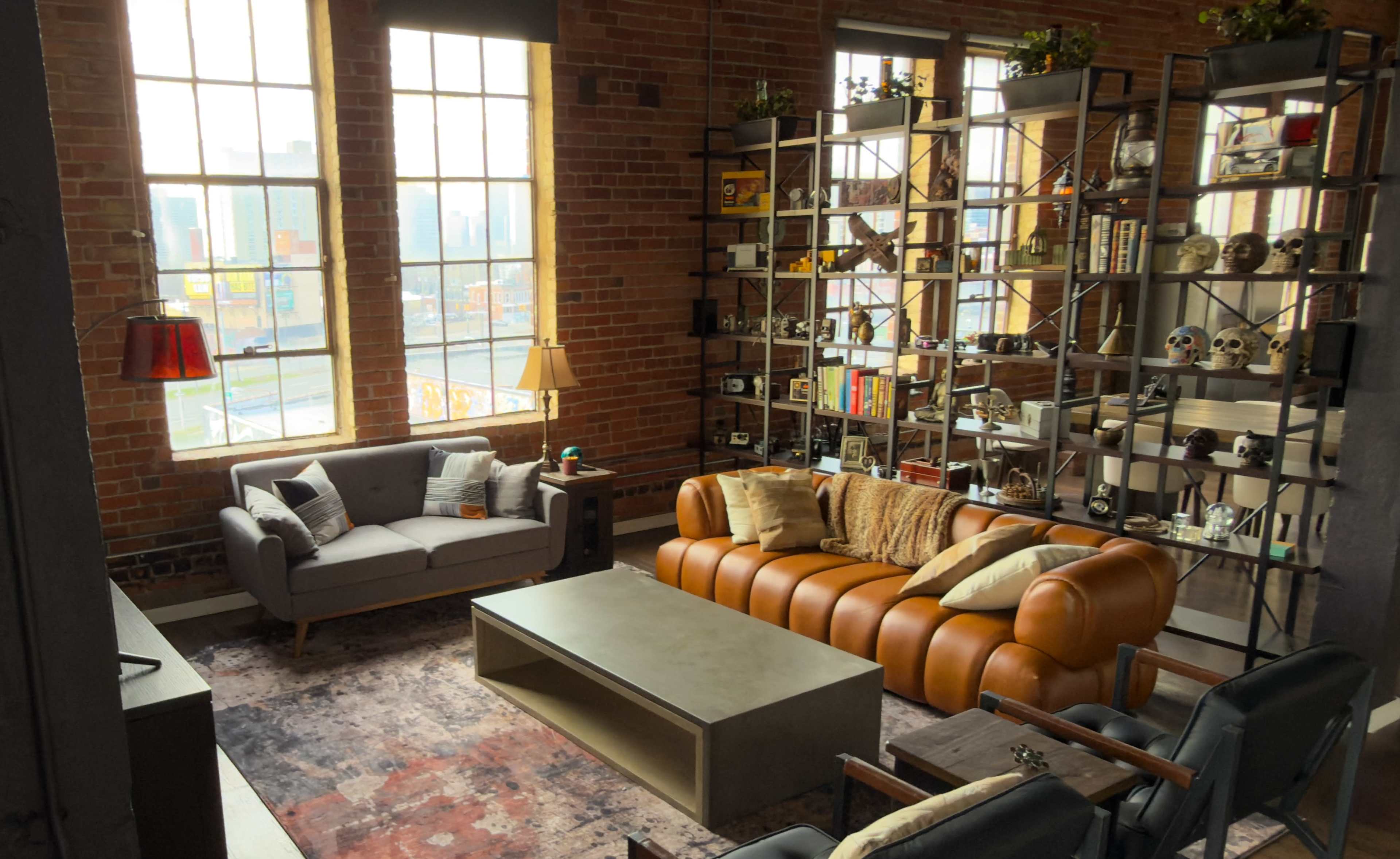 The image shows a cozy living room with large windows, exposed brick walls, a leather sofa, a concrete coffee table, and a bookshelf filled with decorative items.