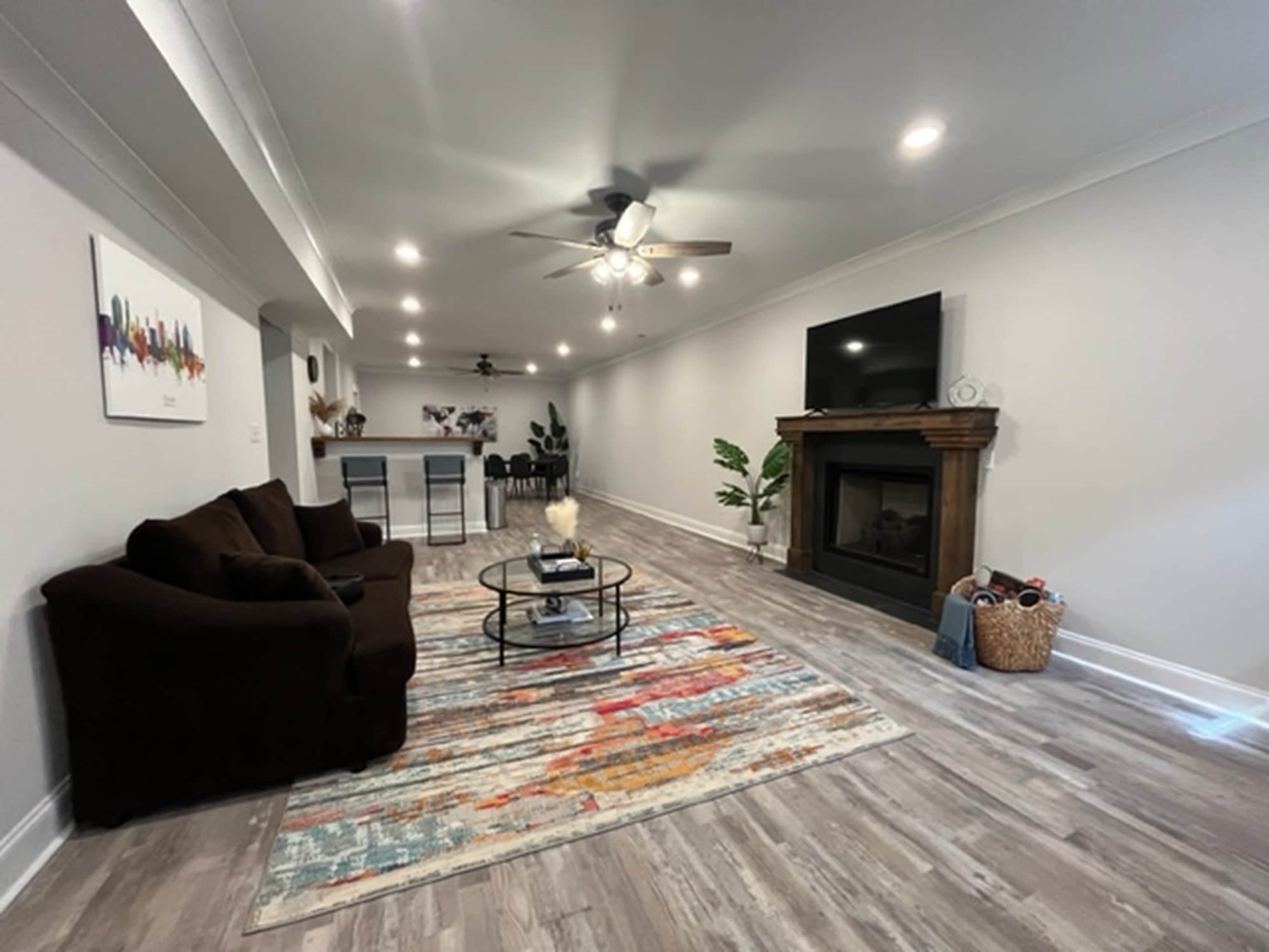 A modern living room with a brown couch, a circular coffee table on a colorful rug, and a wall-mounted TV above a fireplace, leading to a kitchen area in the background.