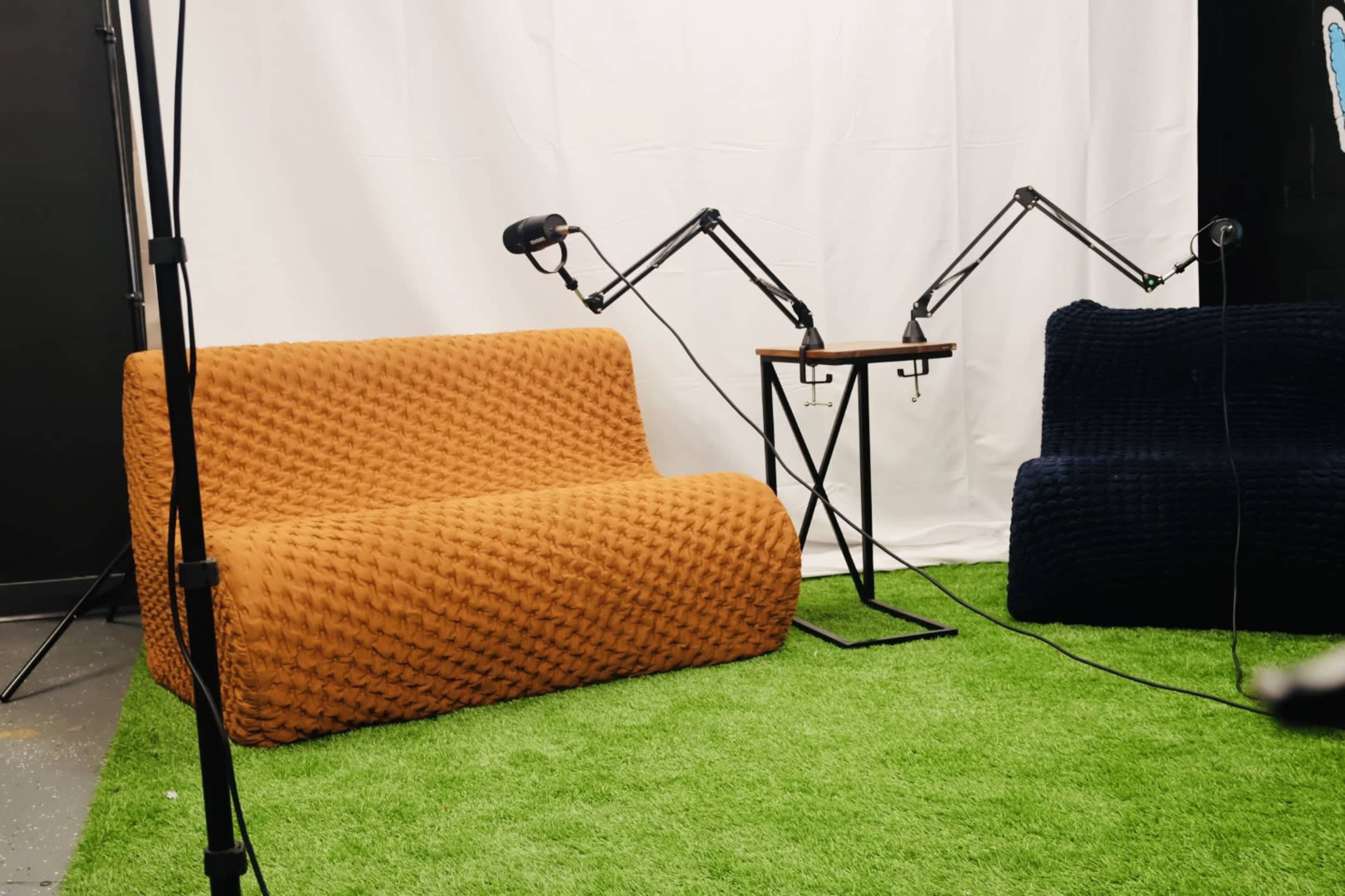 Two sofas, one orange and one navy blue, positioned on a green carpet with a white backdrop and two microphones mounted on stands nearby.
