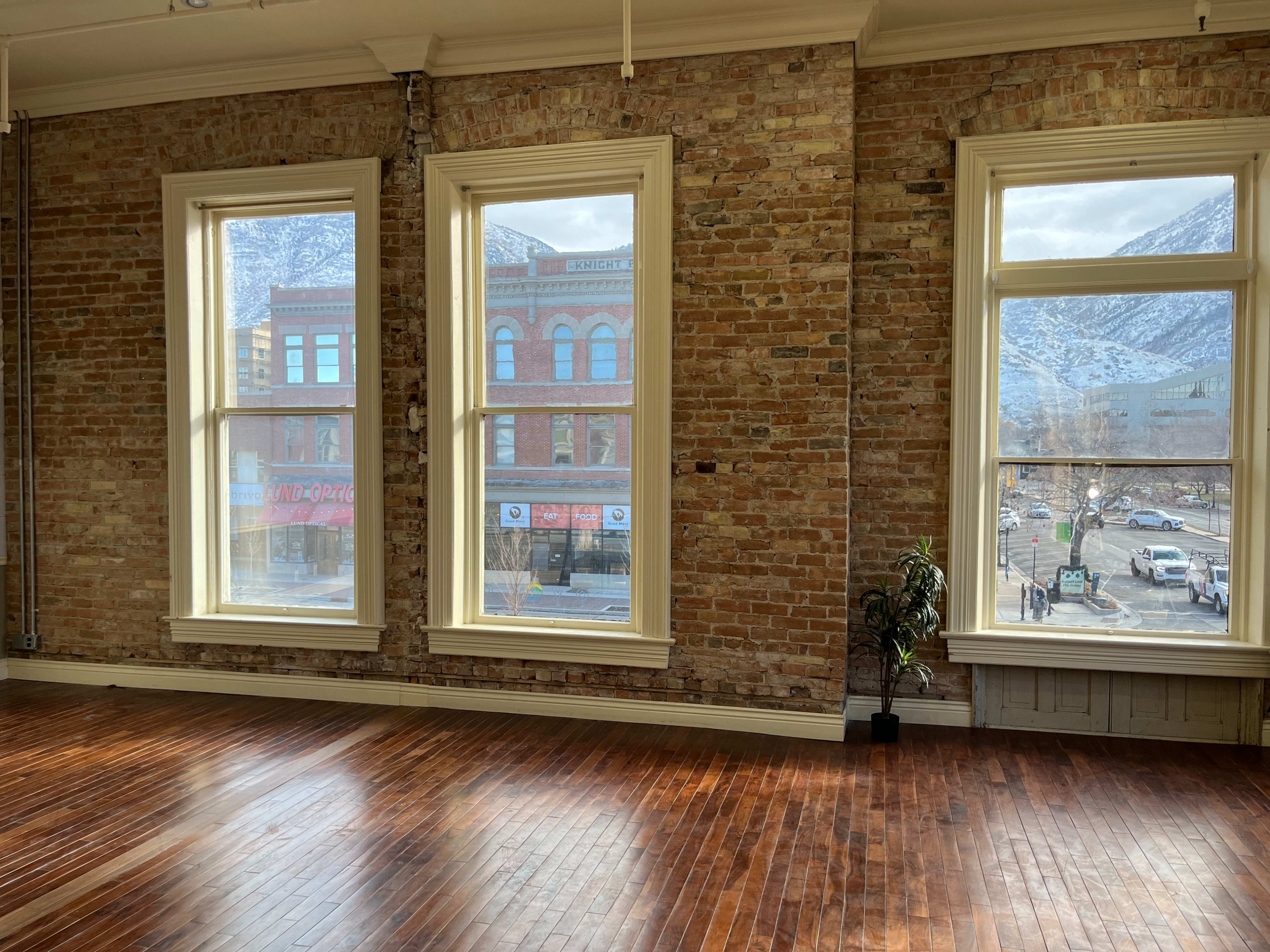 The image shows a spacious room with large windows overlooking a street lined with buildings and mountains in the background.