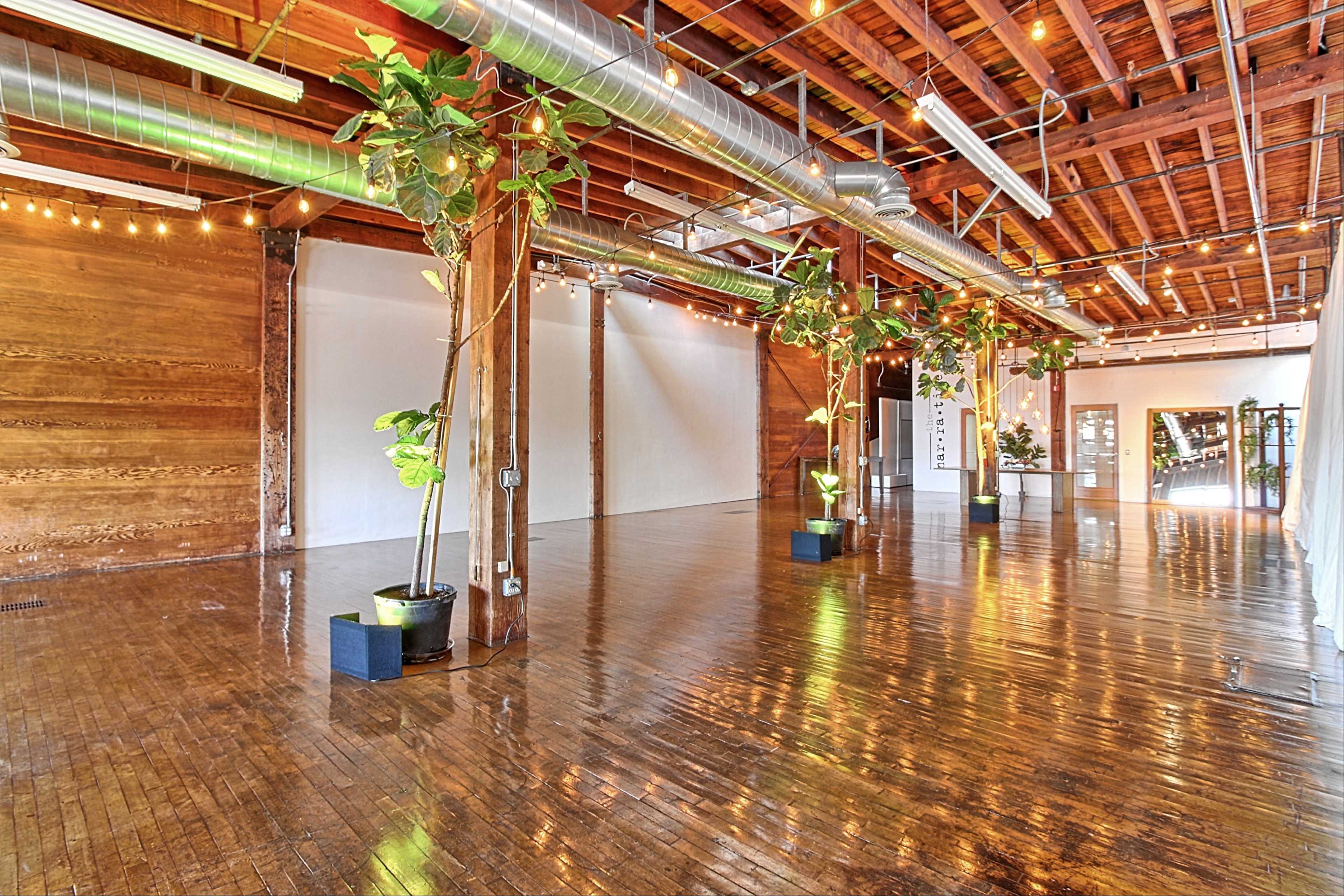 A spacious, hardwood-floored room featuring wooden beams, potted plants, and decorative string lights.