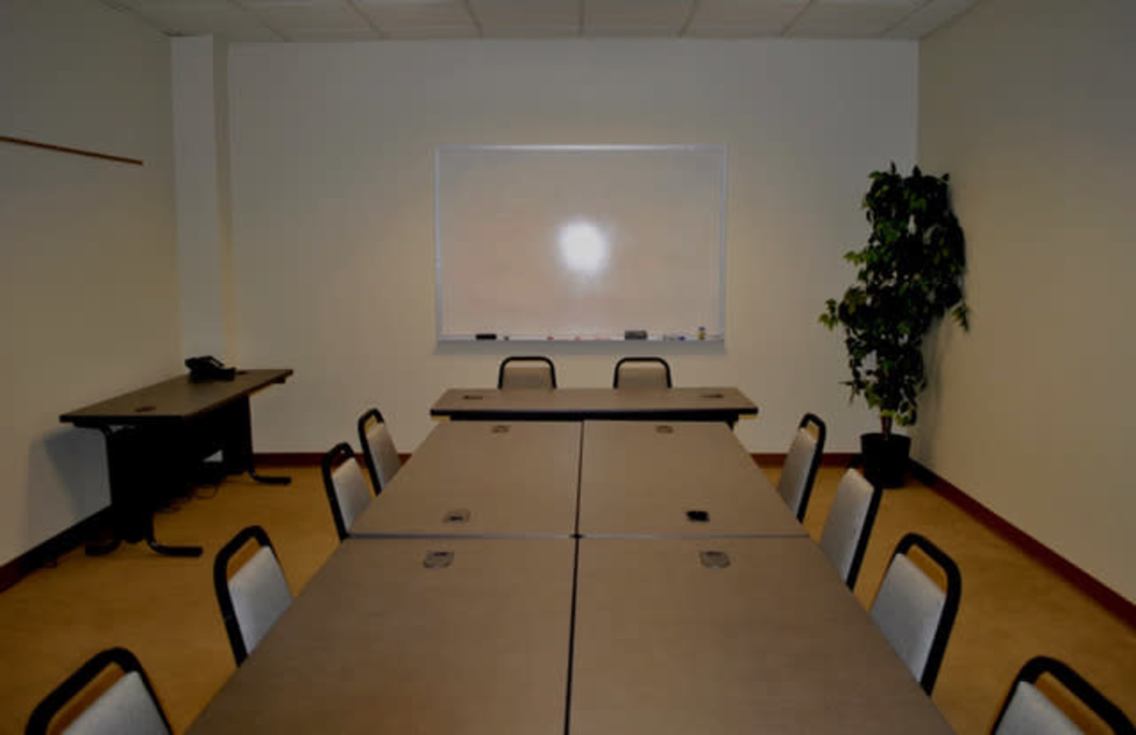 A conference room features several tables arranged in a rectangular formation, with chairs, a whiteboard, and a potted plant in the corner.