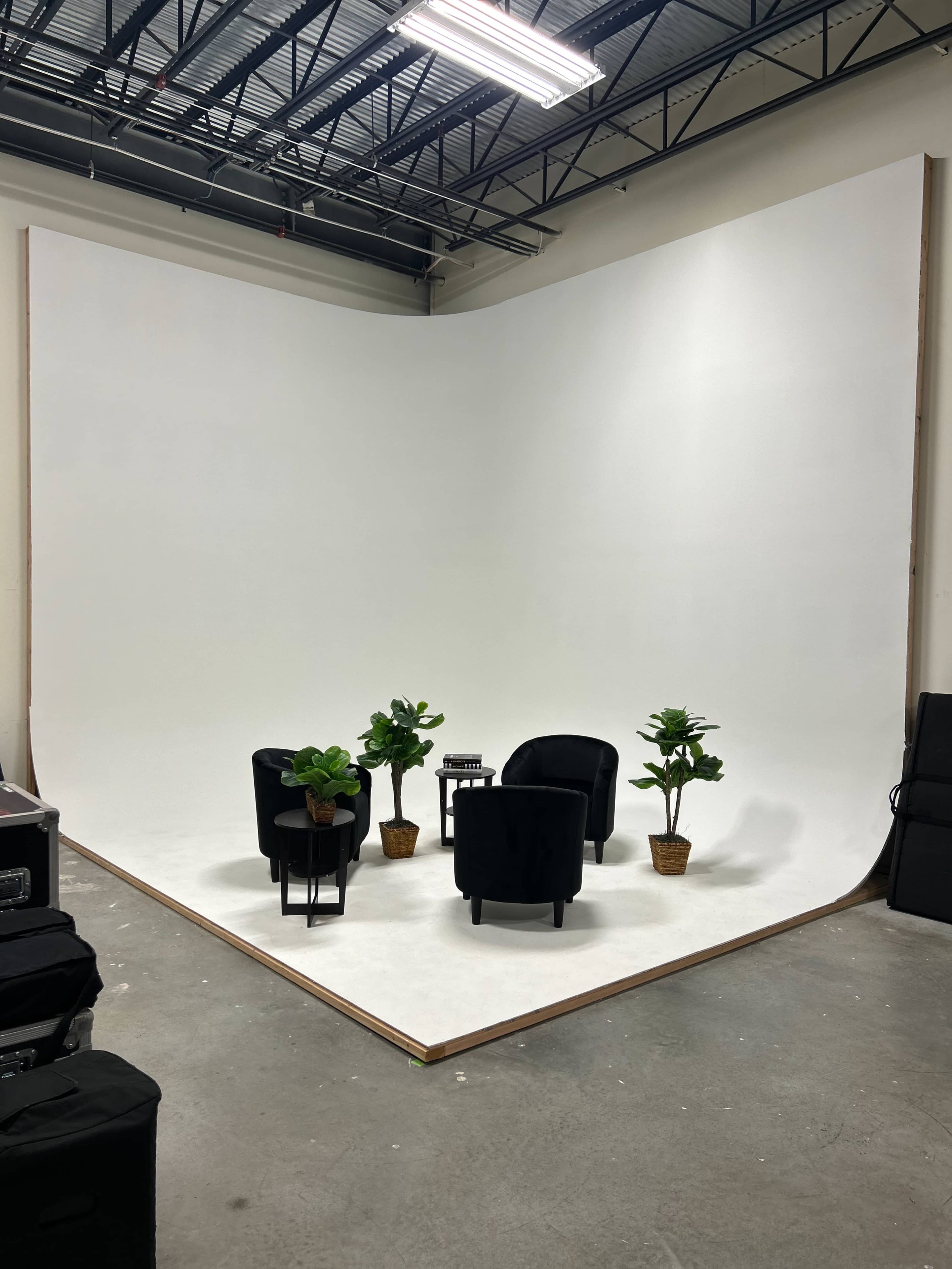 A simple studio setup with two black chairs, a small table, and potted plants against a large, white backdrop.