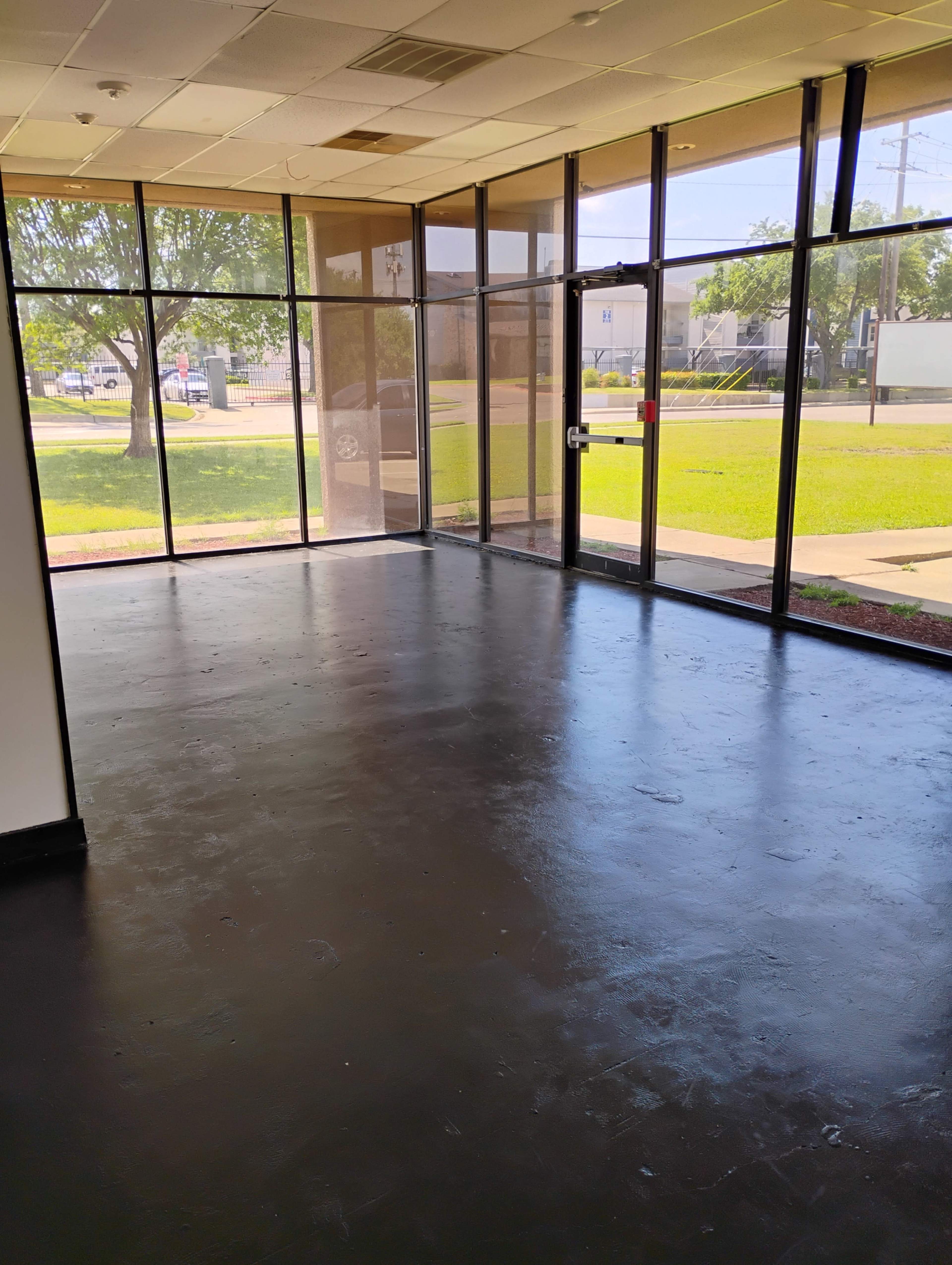 The image shows an empty commercial space with a black floor and large glass windows allowing natural light to enter.