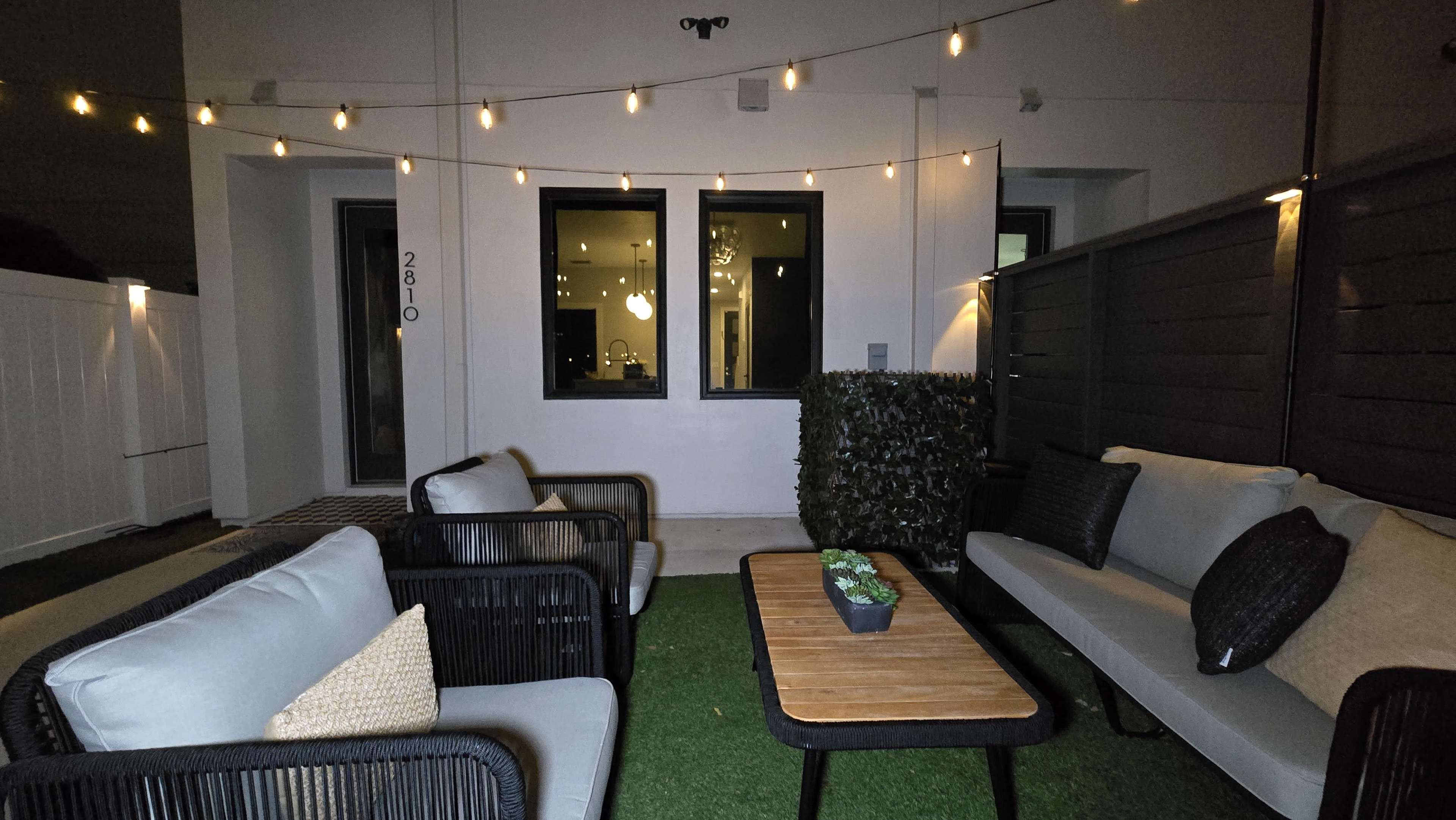 The image shows an outdoor seating area with two couches, a wooden coffee table, and decorative lights overhead, flanked by a hedge and a wall.