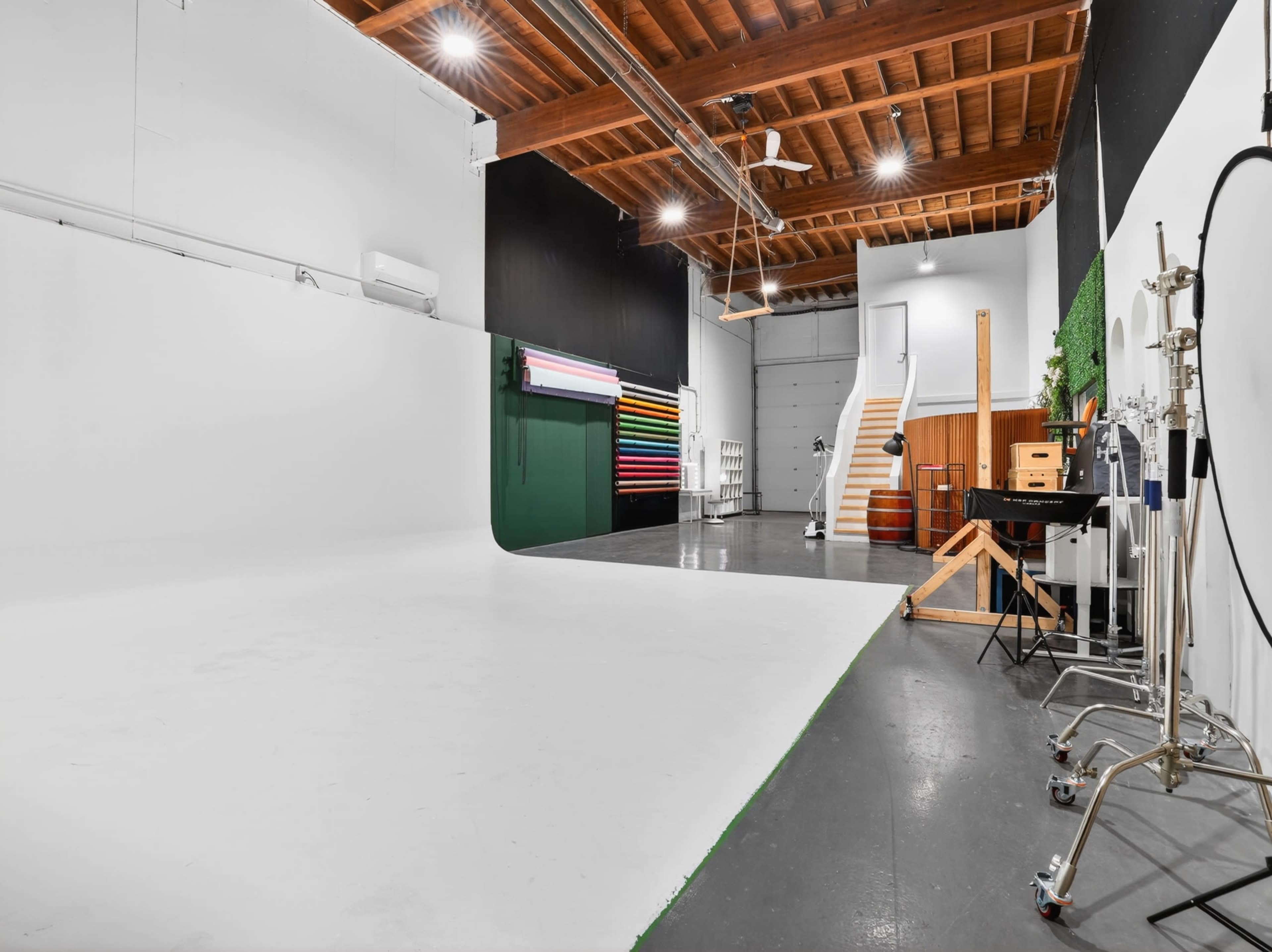 The image shows a photography studio featuring a large white backdrop, colorful lighting equipment, and various studio props against a wooden ceiling.