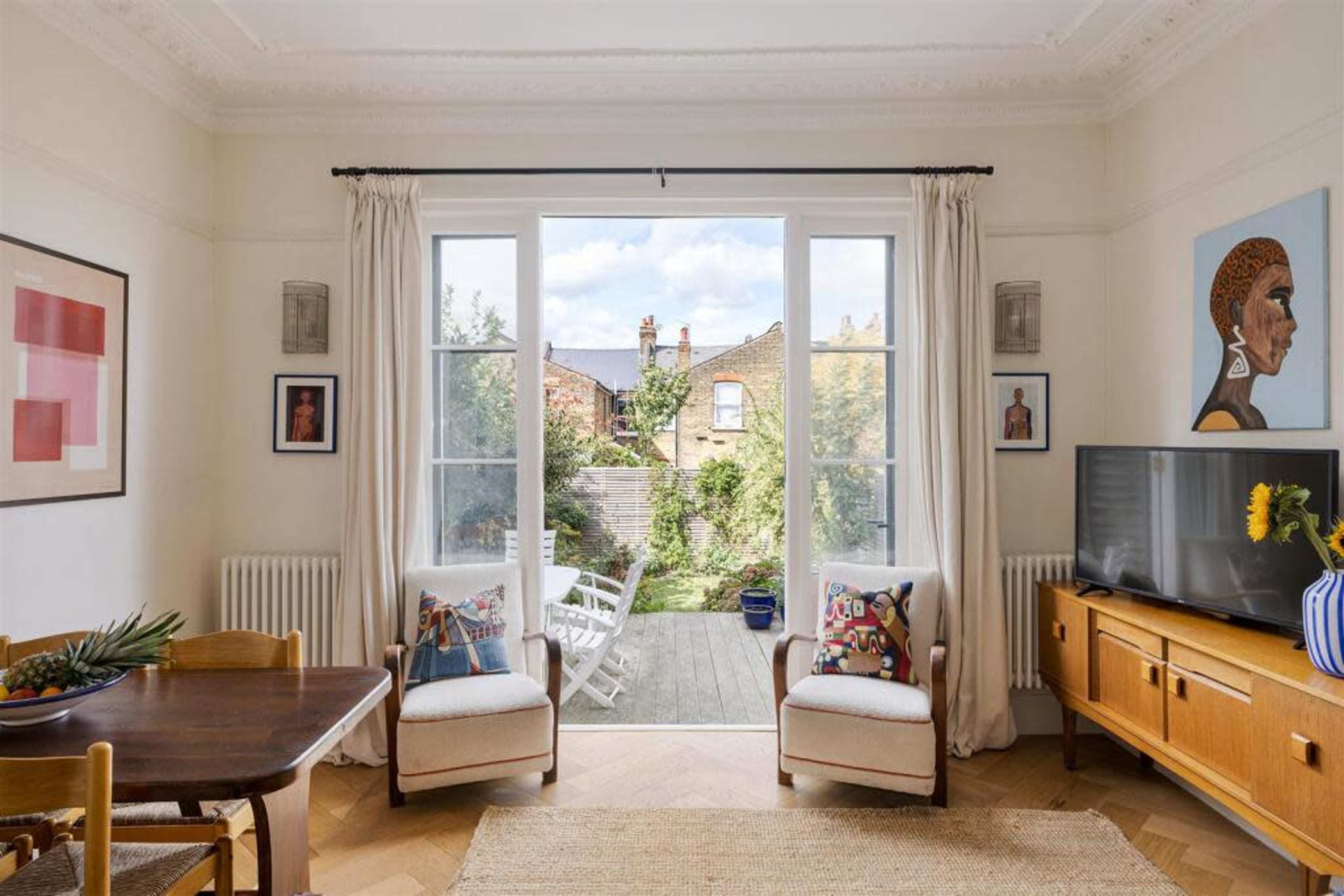 The image shows a bright living room with large windows overlooking a garden, featuring a dining table, two armchairs, a television, and various wall art.