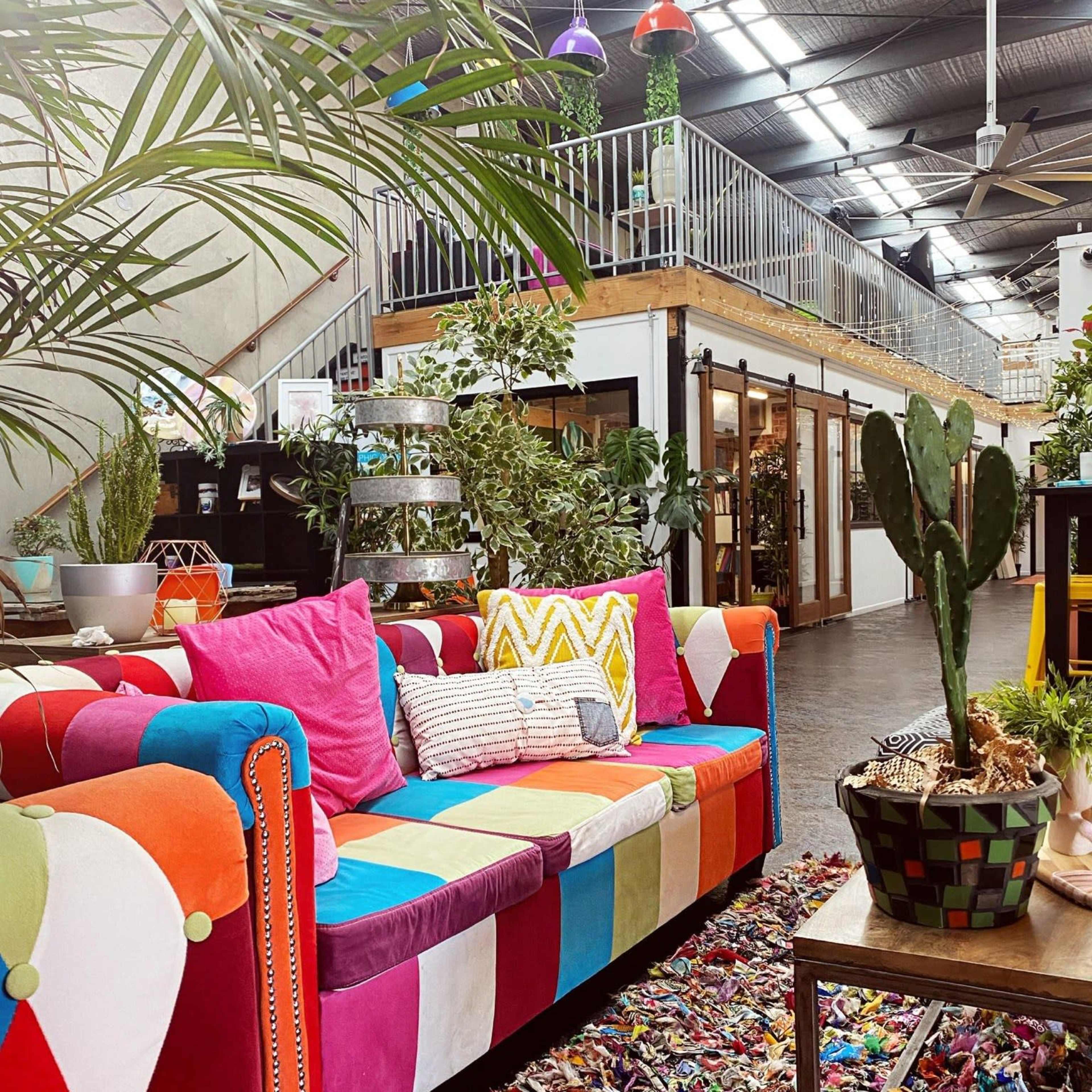 A brightly colored patchwork sofa sits in a lush, plant-filled indoor space with an upper level and decorative lights.
