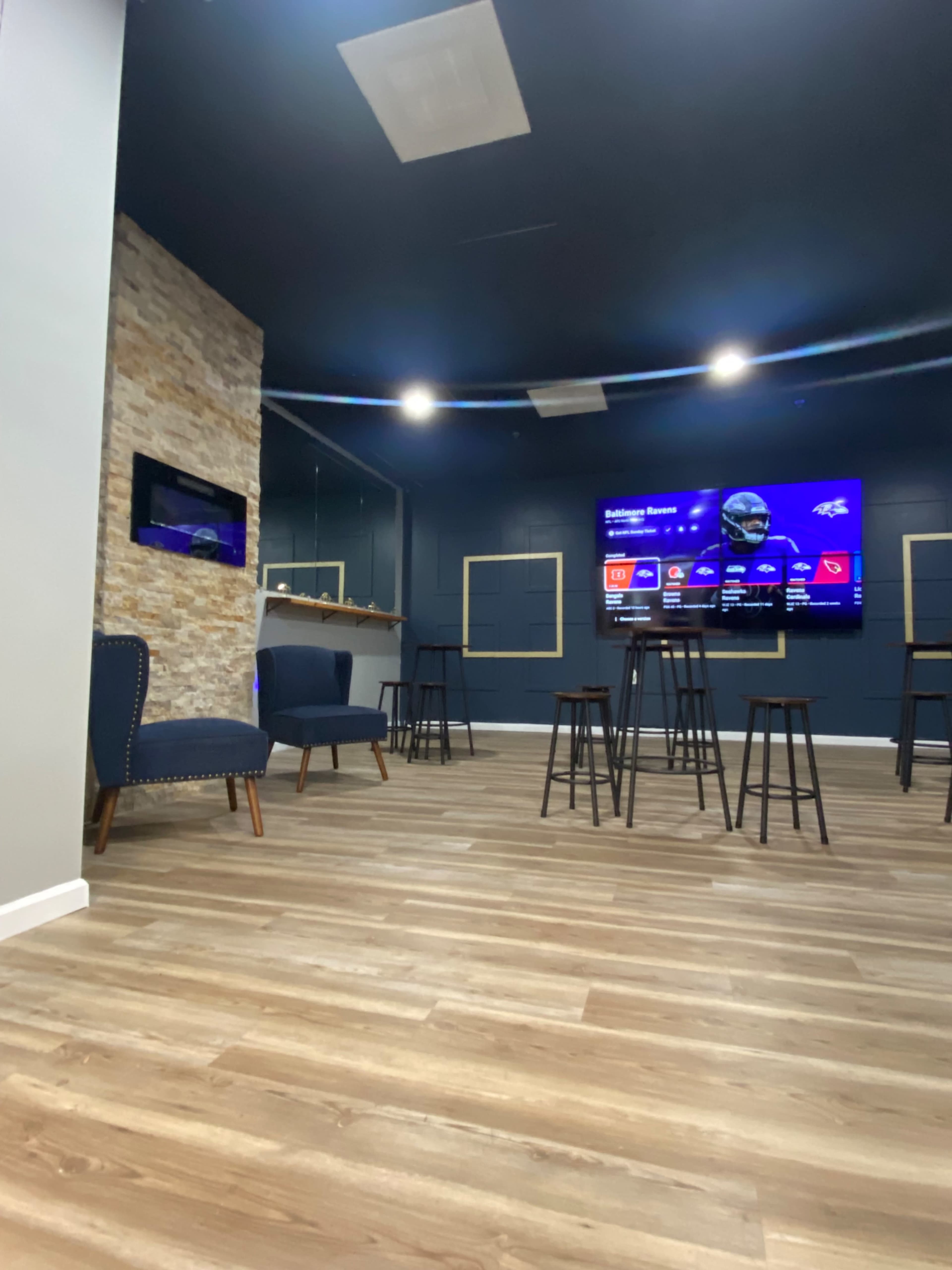 The image shows a modern lounge area with wooden flooring, two blue chairs, and several tall black stools arranged in front of a large screen displaying information on a wall.