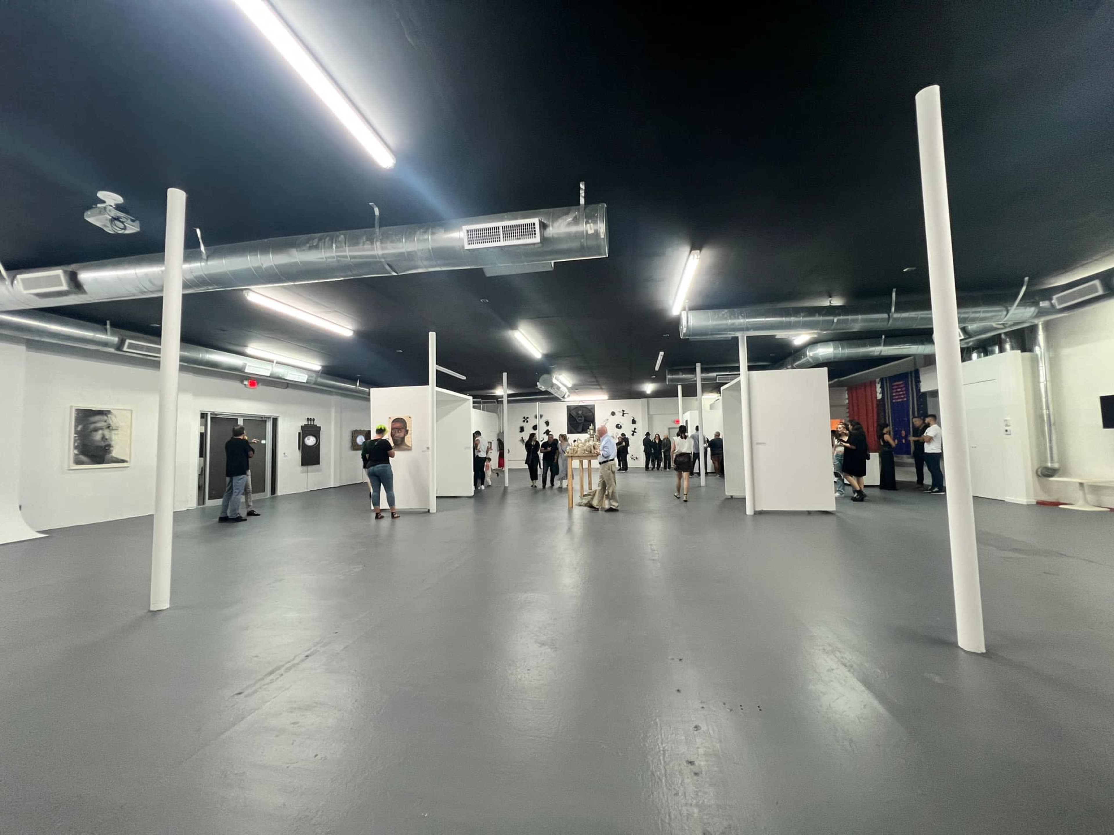 A spacious art gallery features white walls and a gray floor, filled with visitors examining the displayed works.