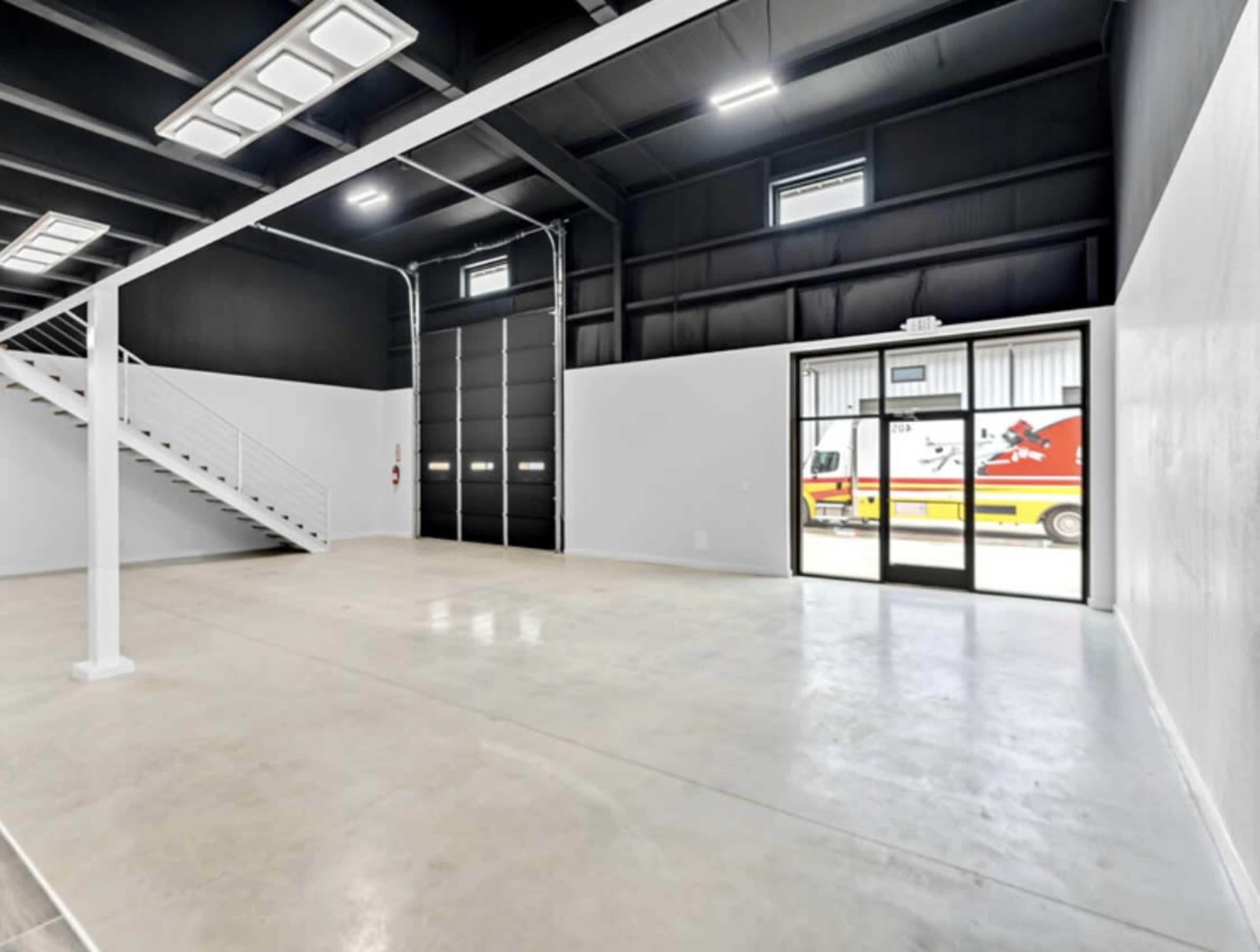 The image shows a spacious, empty interior of a warehouse with a staircase, large windows, and a vehicle visible through the glass doors.
