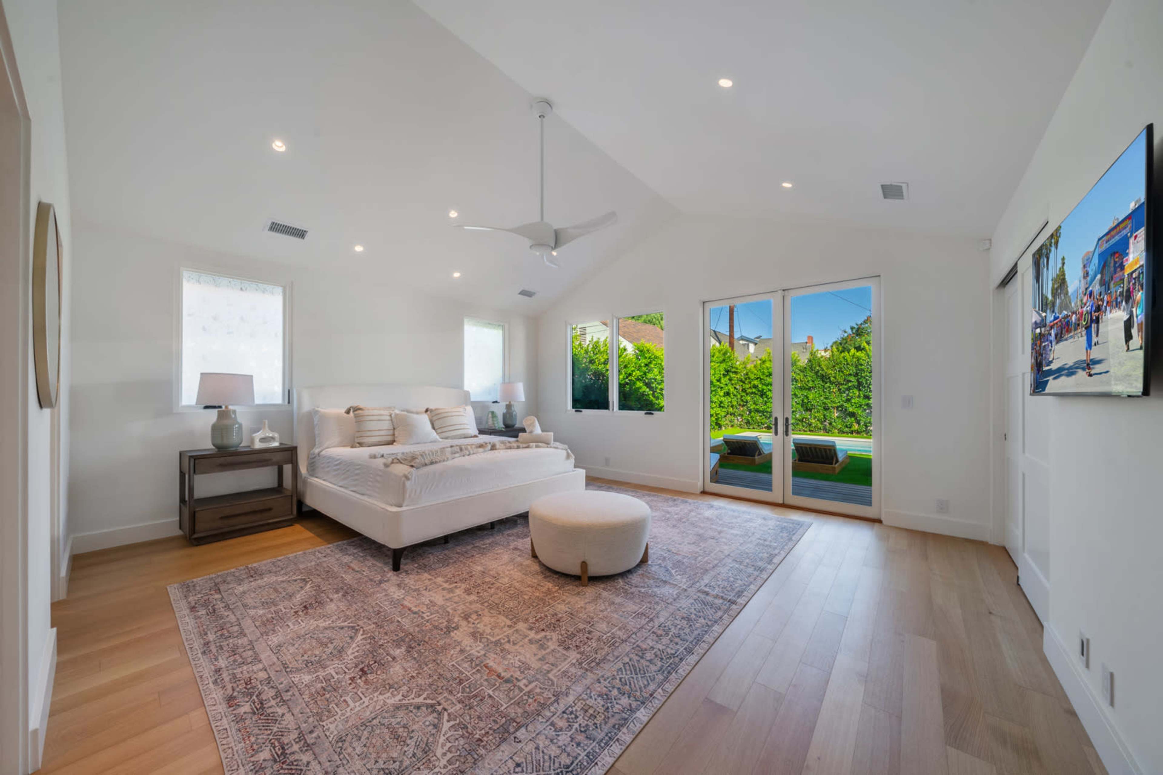 The image shows a spacious, modern bedroom featuring a large bed, a round ottoman, a wooden nightstand, and glass doors leading to an outdoor area.