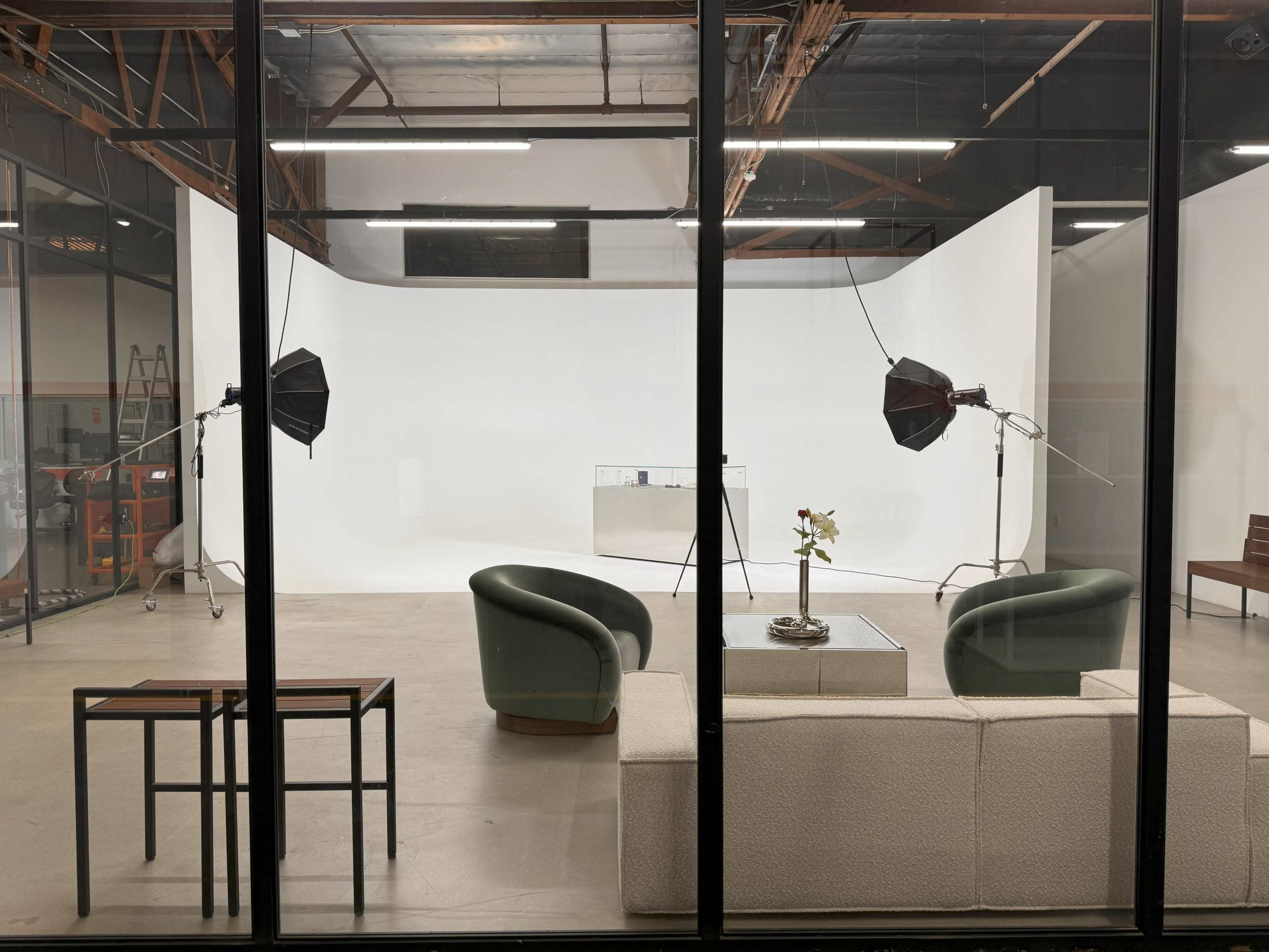 The image shows a modern studio space featuring two distinct seating areas, a table, and professional lighting equipment set up in front of a white backdrop.