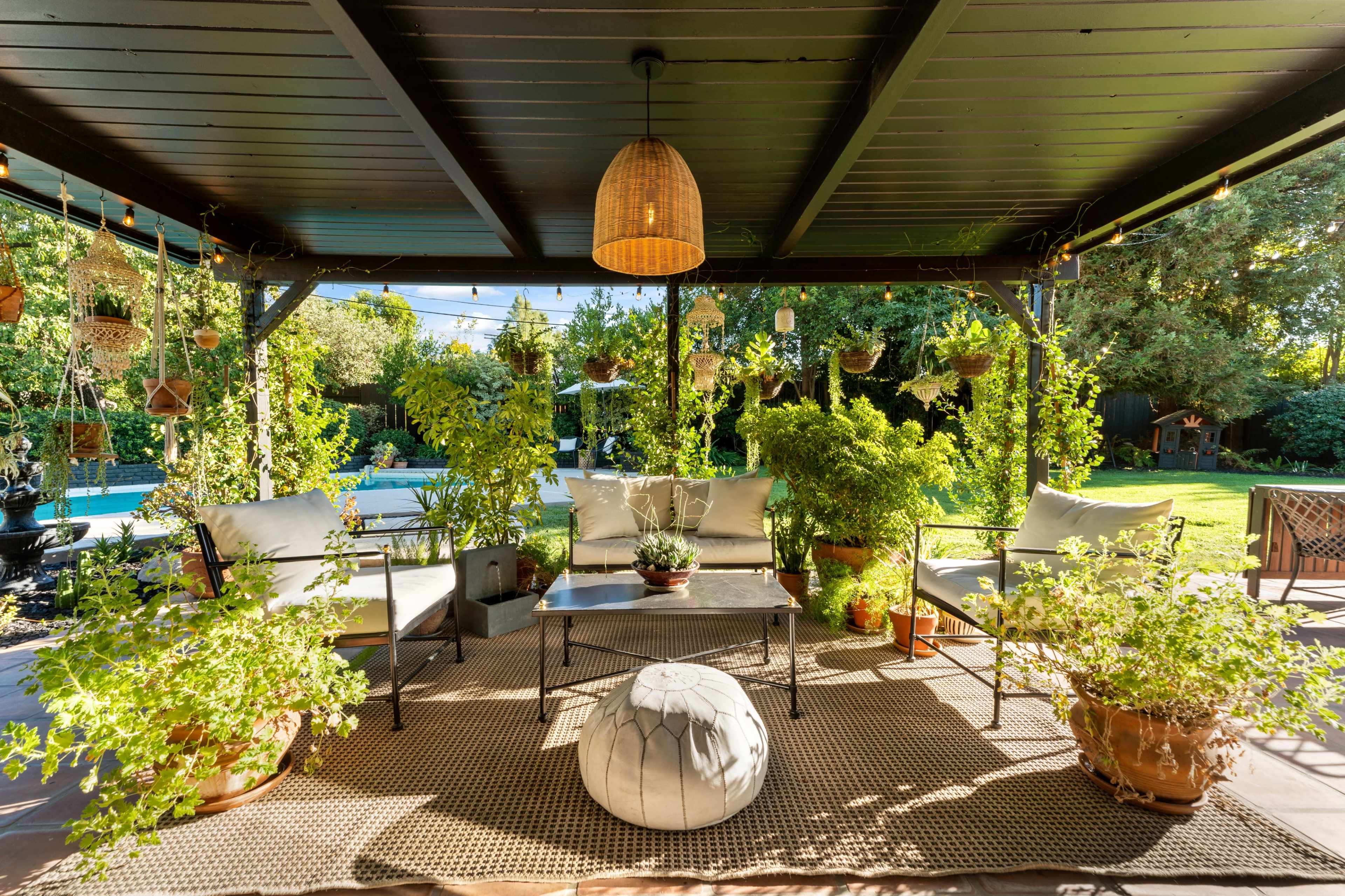 A covered patio area with seating arrangements, potted plants, and decorative lighting surrounded by a lush garden.