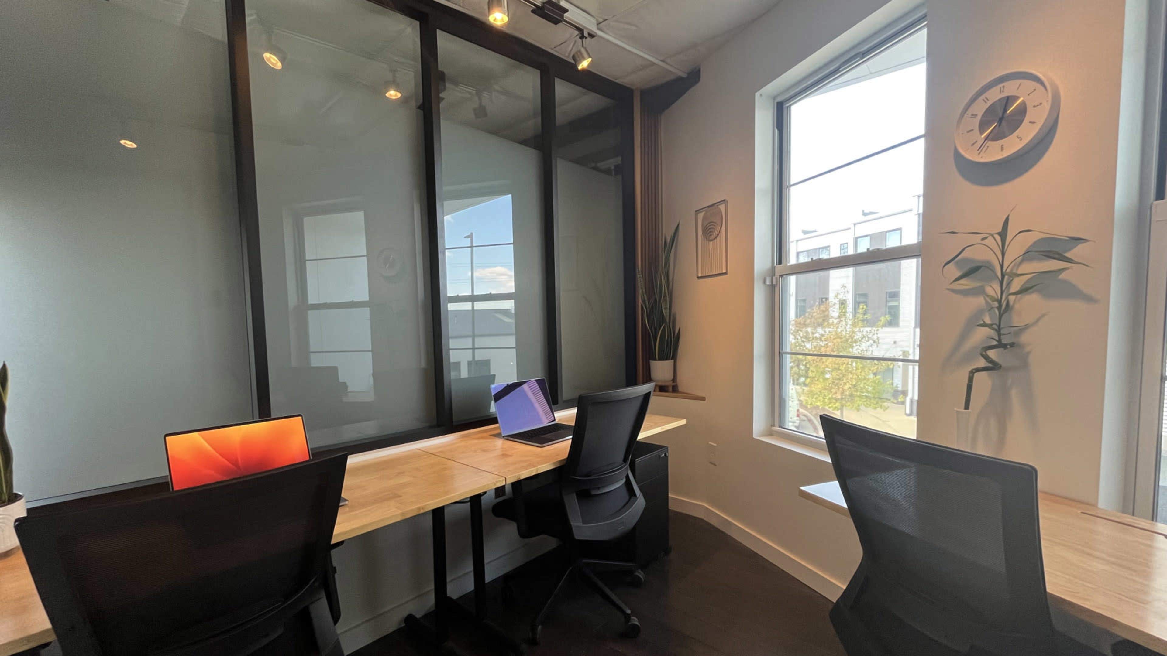 The image shows a modern office space with two desks, each equipped with a laptop, against a backdrop of large windows and partitioned glass walls.