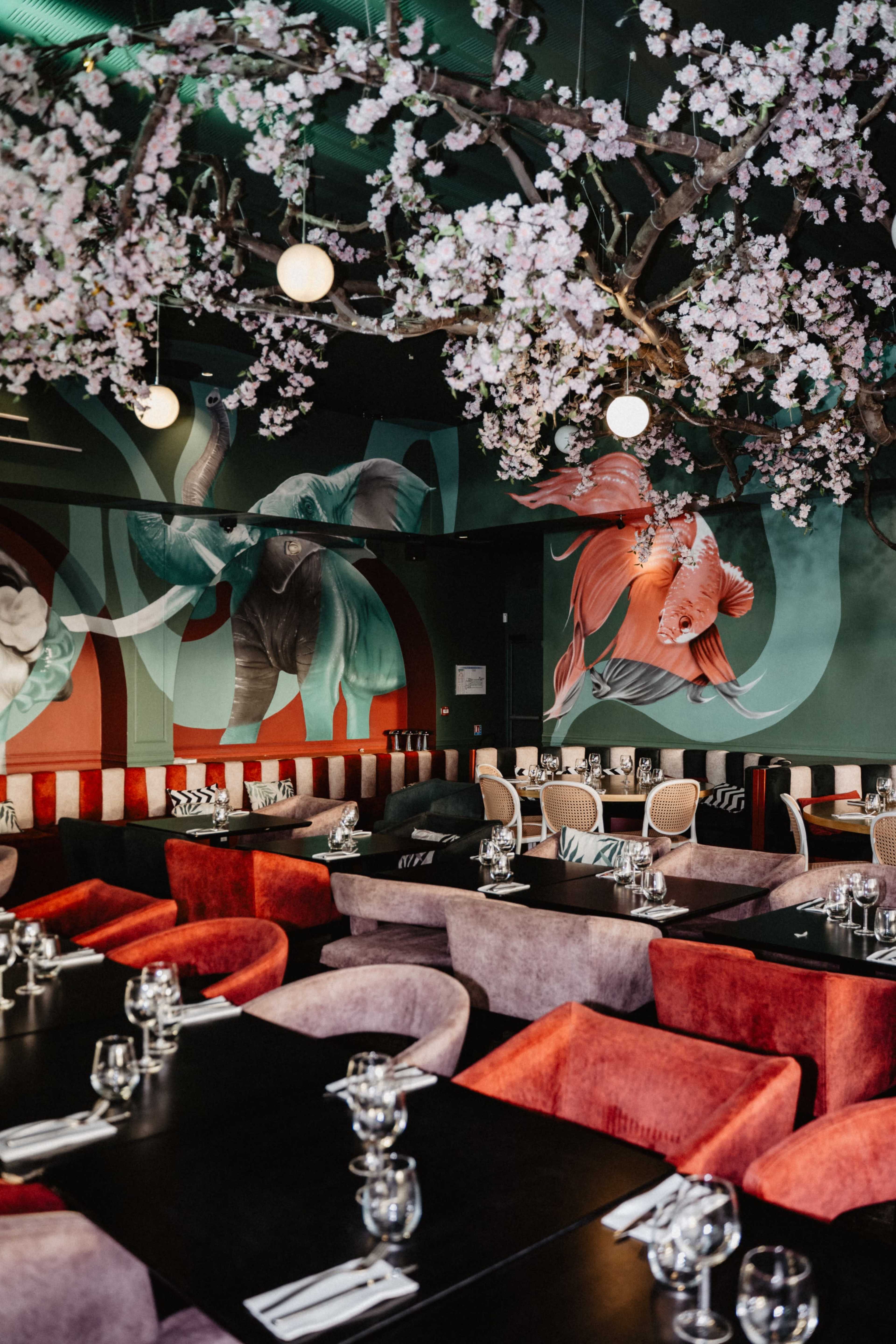 The image shows a restaurant interior featuring large murals of elephants and fish, with cherry blossom decorations hanging from the ceiling above tables set for dining.