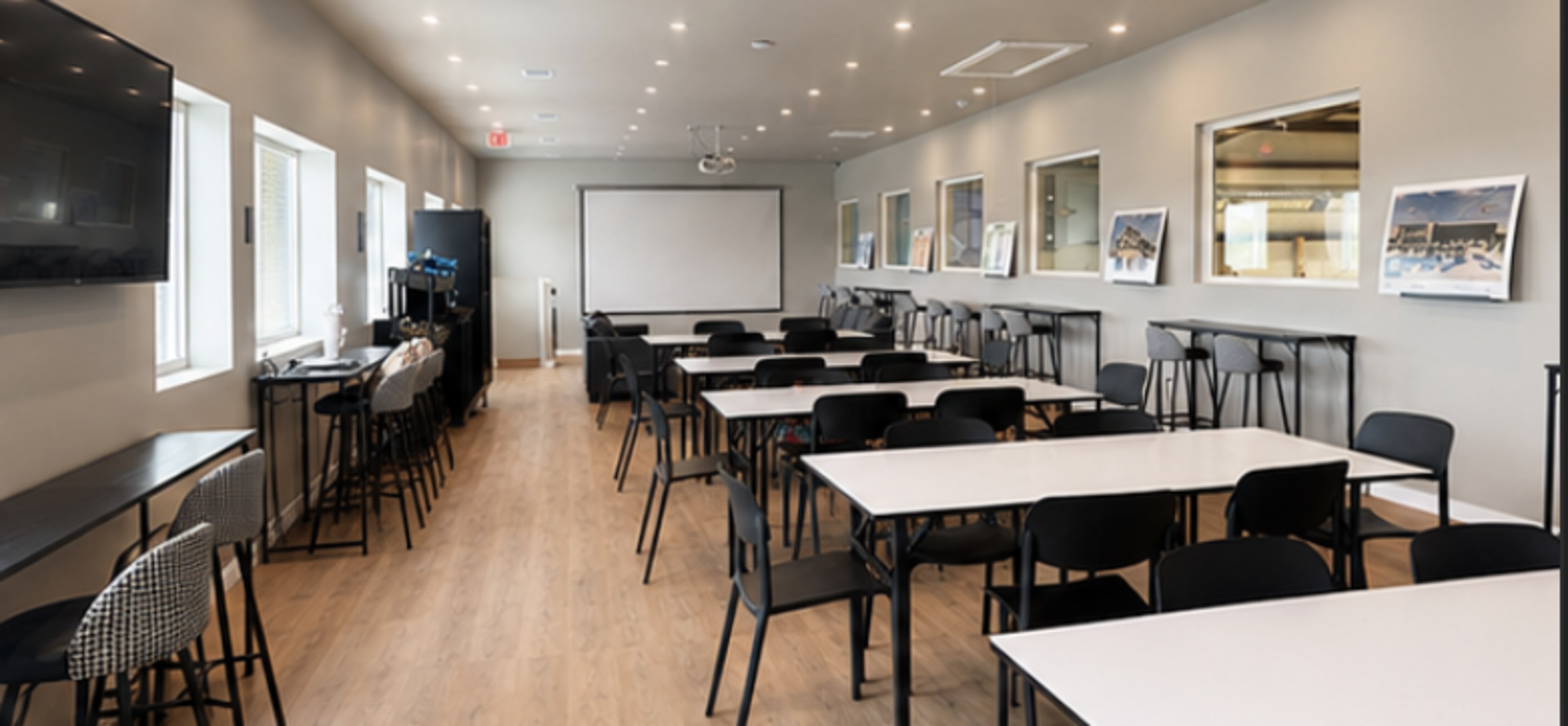 The image shows a meeting room with rows of black chairs and white tables arranged in a spacious layout, featuring large windows and a presentation screen.