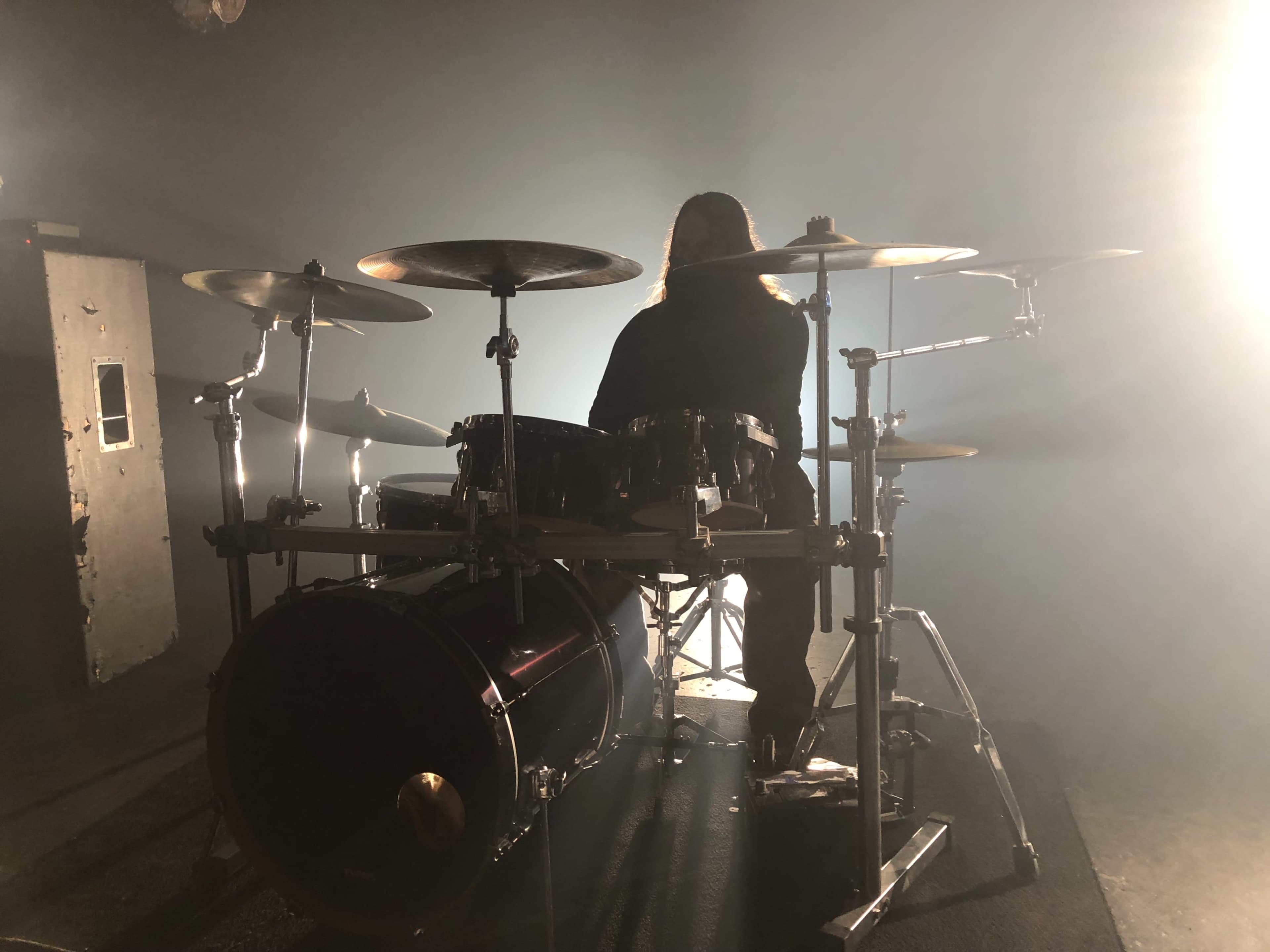 A drummer sits behind a drum set in a dimly lit space with beams of light illuminating the scene.