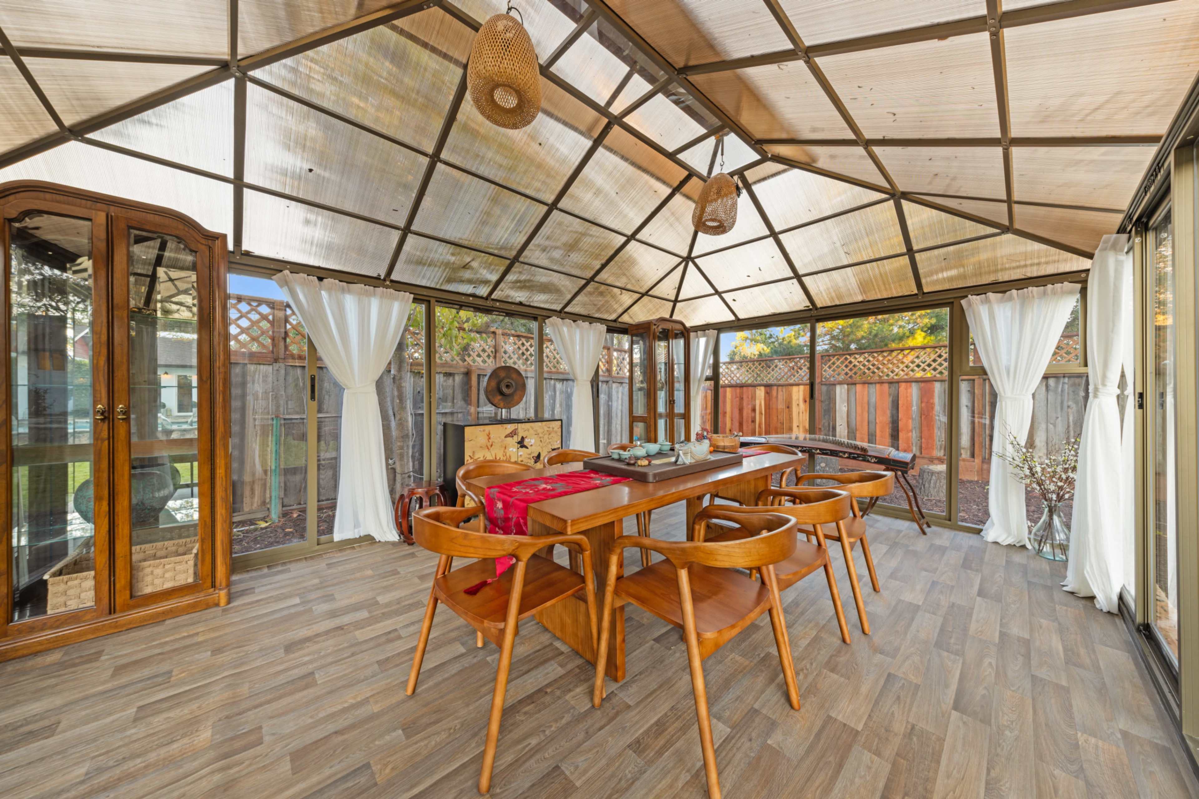 The image shows a sunroom or enclosed patio featuring a wooden table surrounded by chairs, with large windows and curtains, and decorative elements like woven light fixtures and a display cabinet.