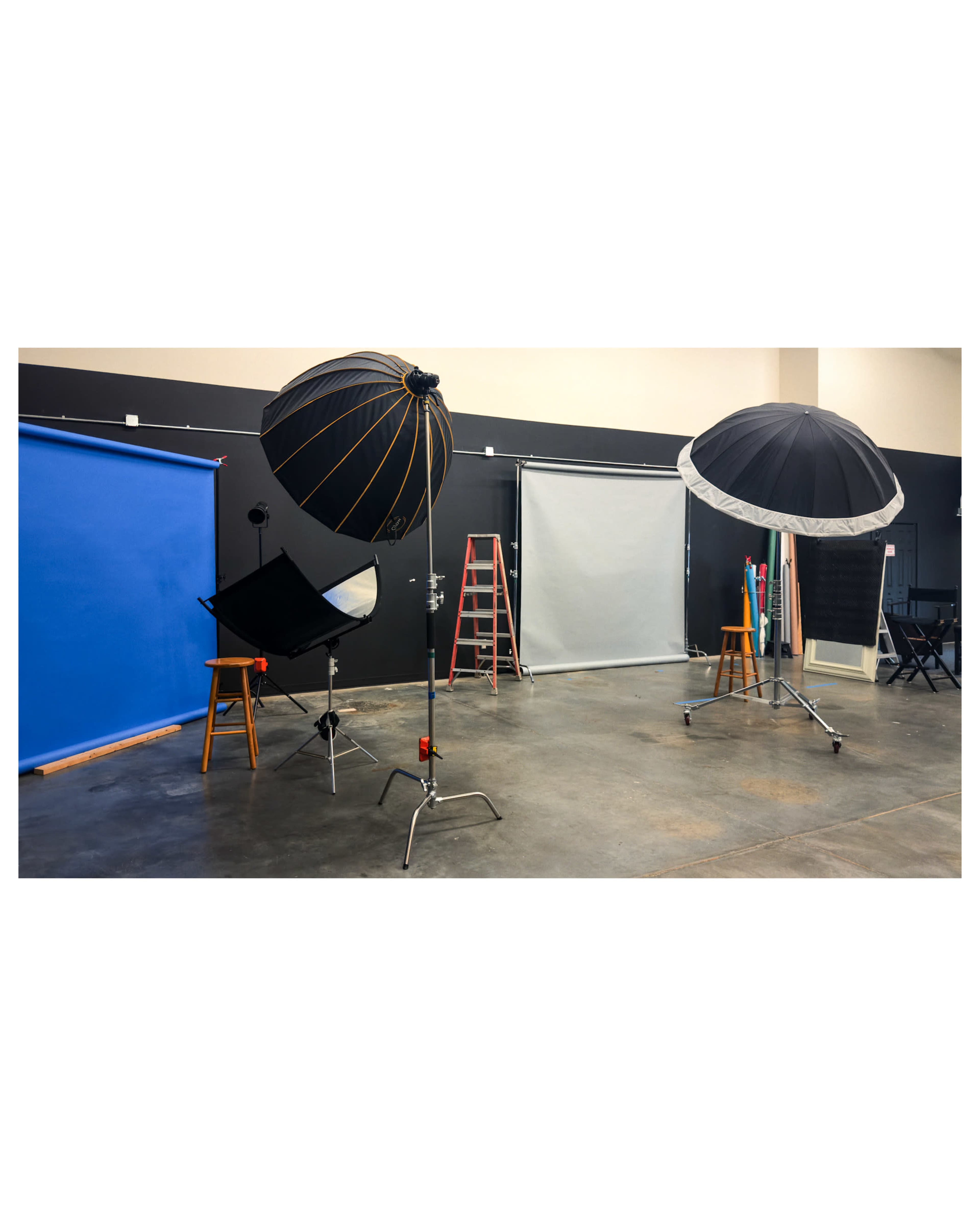 The image shows a photography studio with lighting equipment, backdrops, and a stepladder arranged around the space.