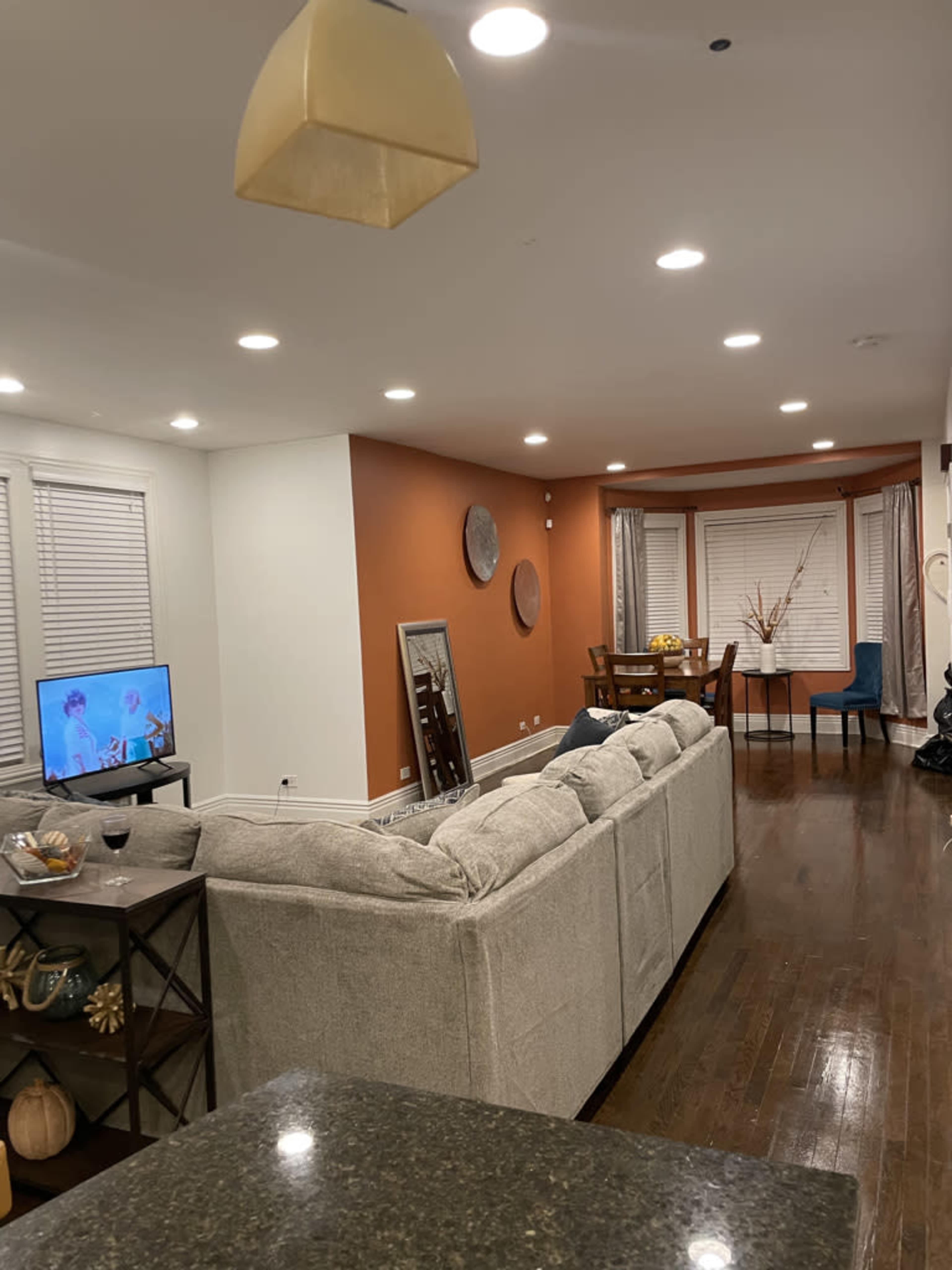 A living room features a gray sofa facing a television, with an adjacent dining area and warm-colored accent wall.