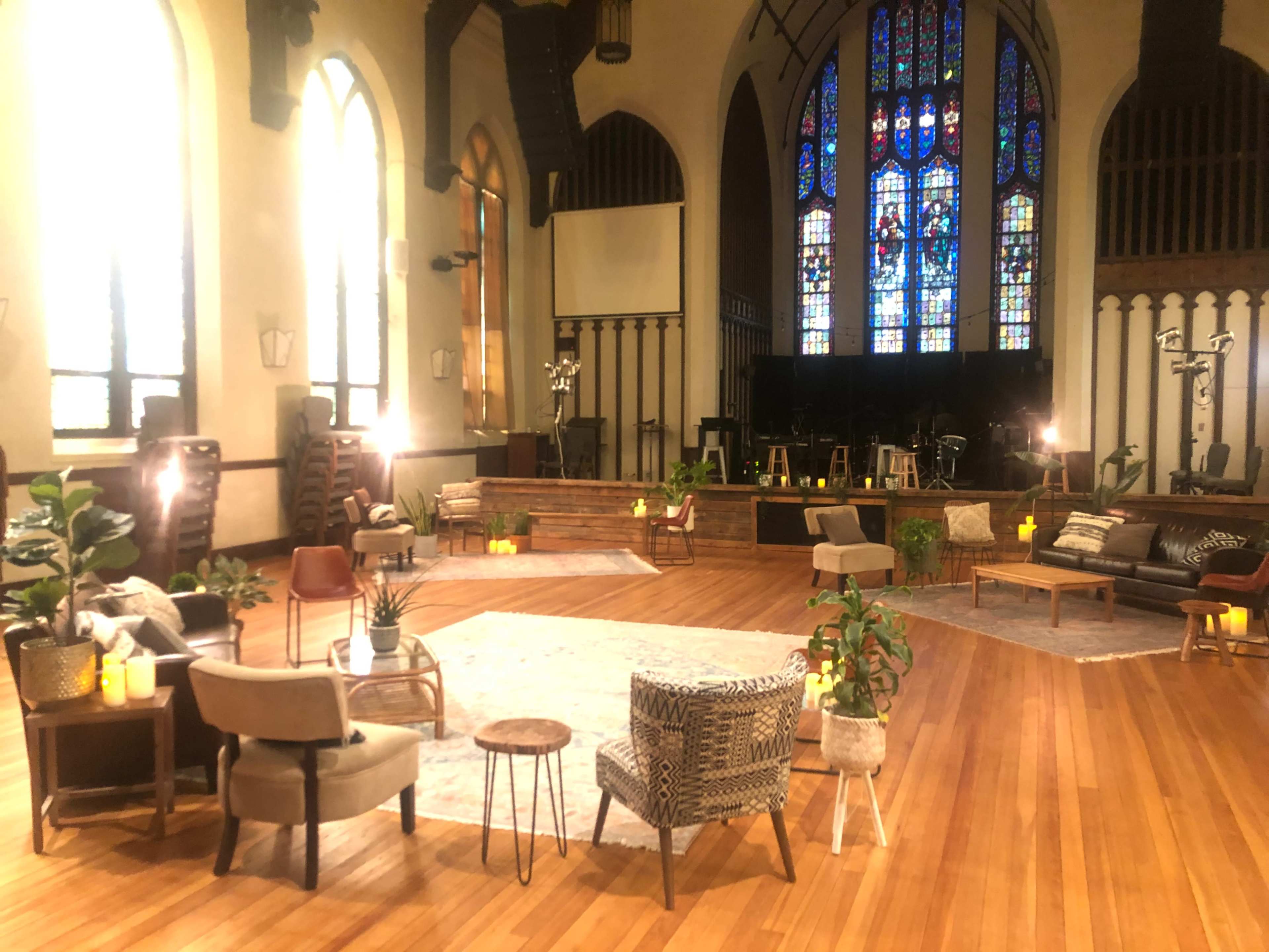 A spacious interior of a church features a circular arrangement of various seating options and decorative plants, set against colorful stained glass windows.