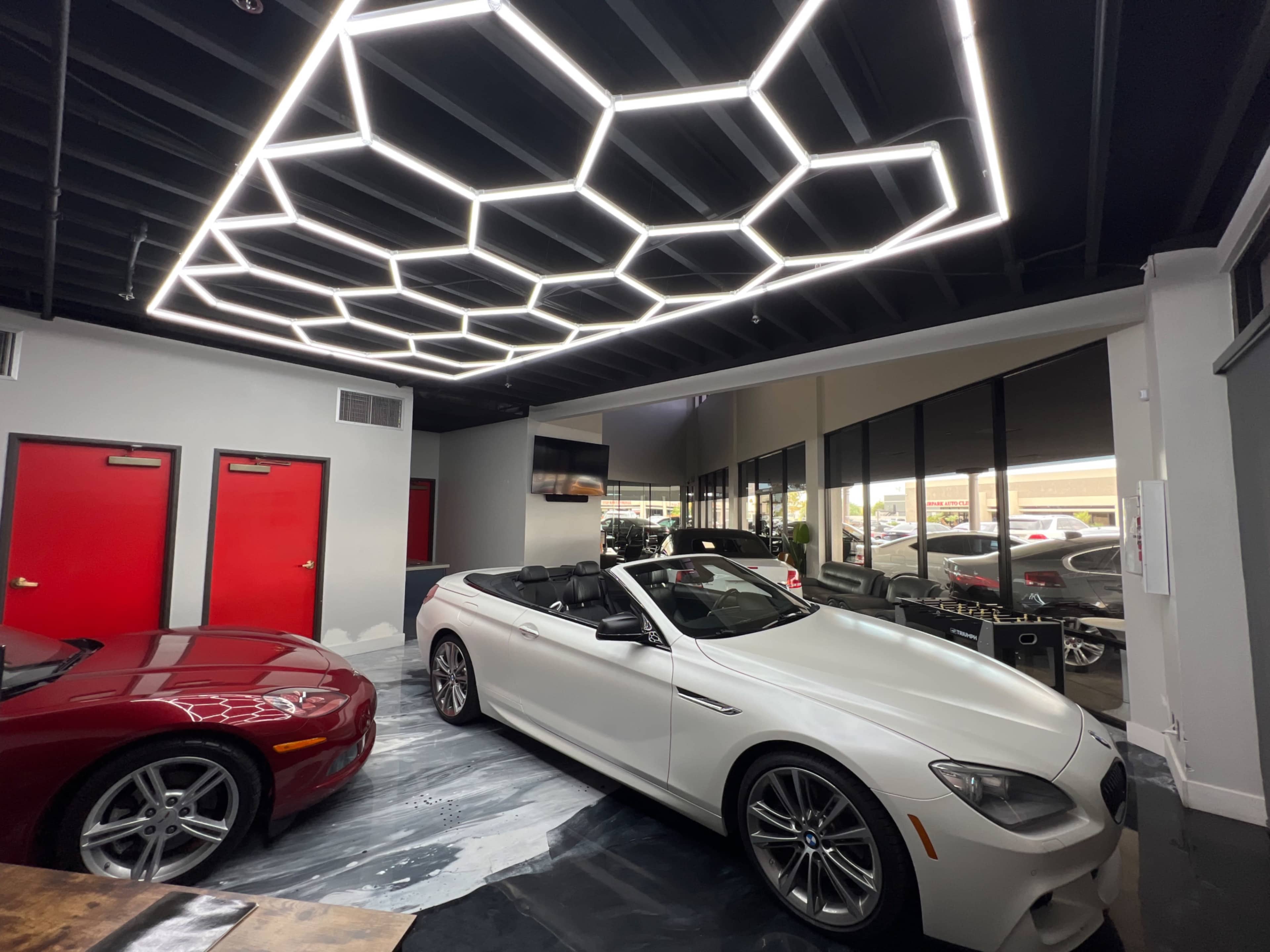 A sleek white convertible car parked in a modern showroom featuring a honeycomb-patterned light fixture and red doors.