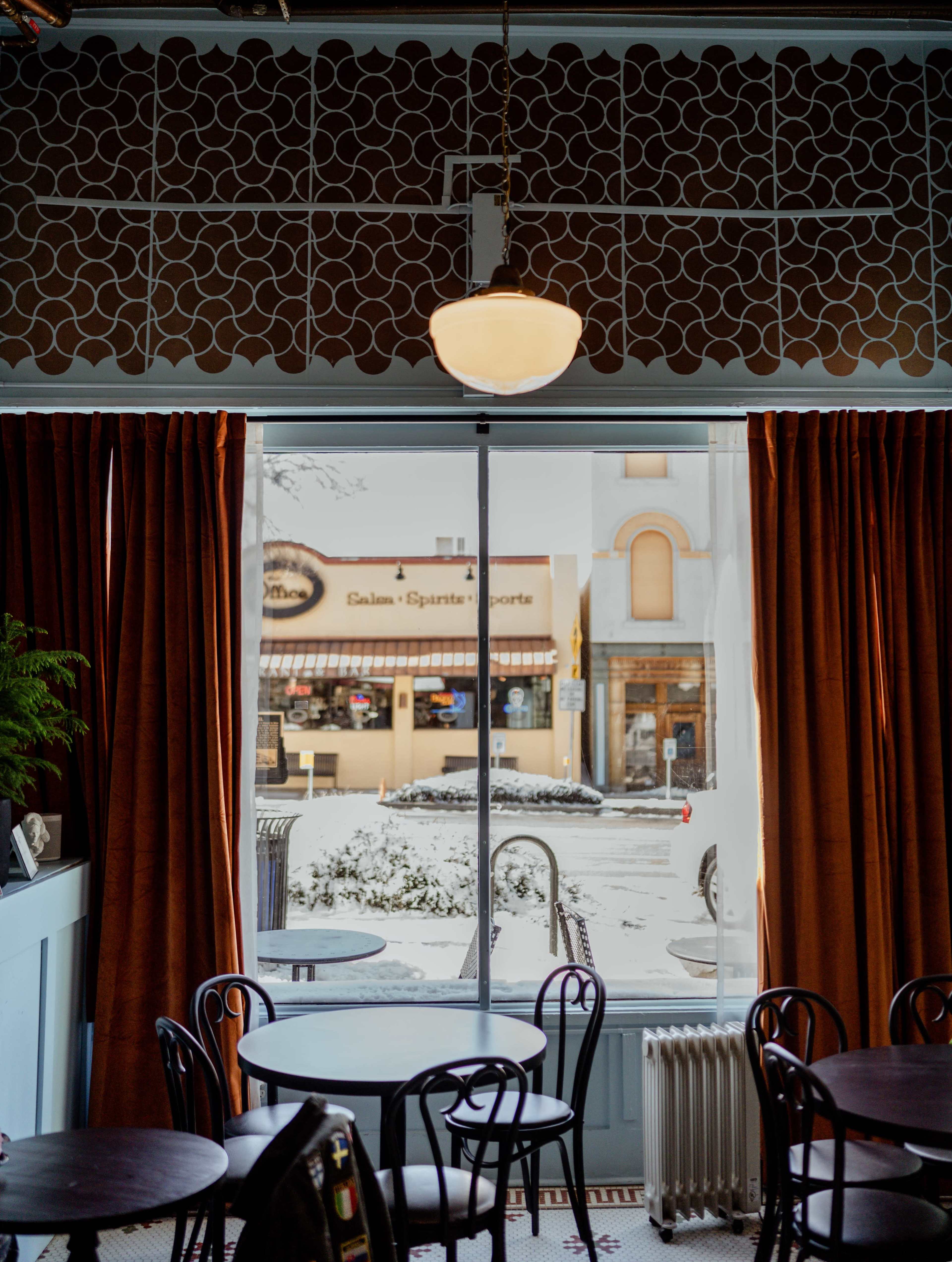 A cozy café interior features round tables and brown curtains framing a snowy street view outside.