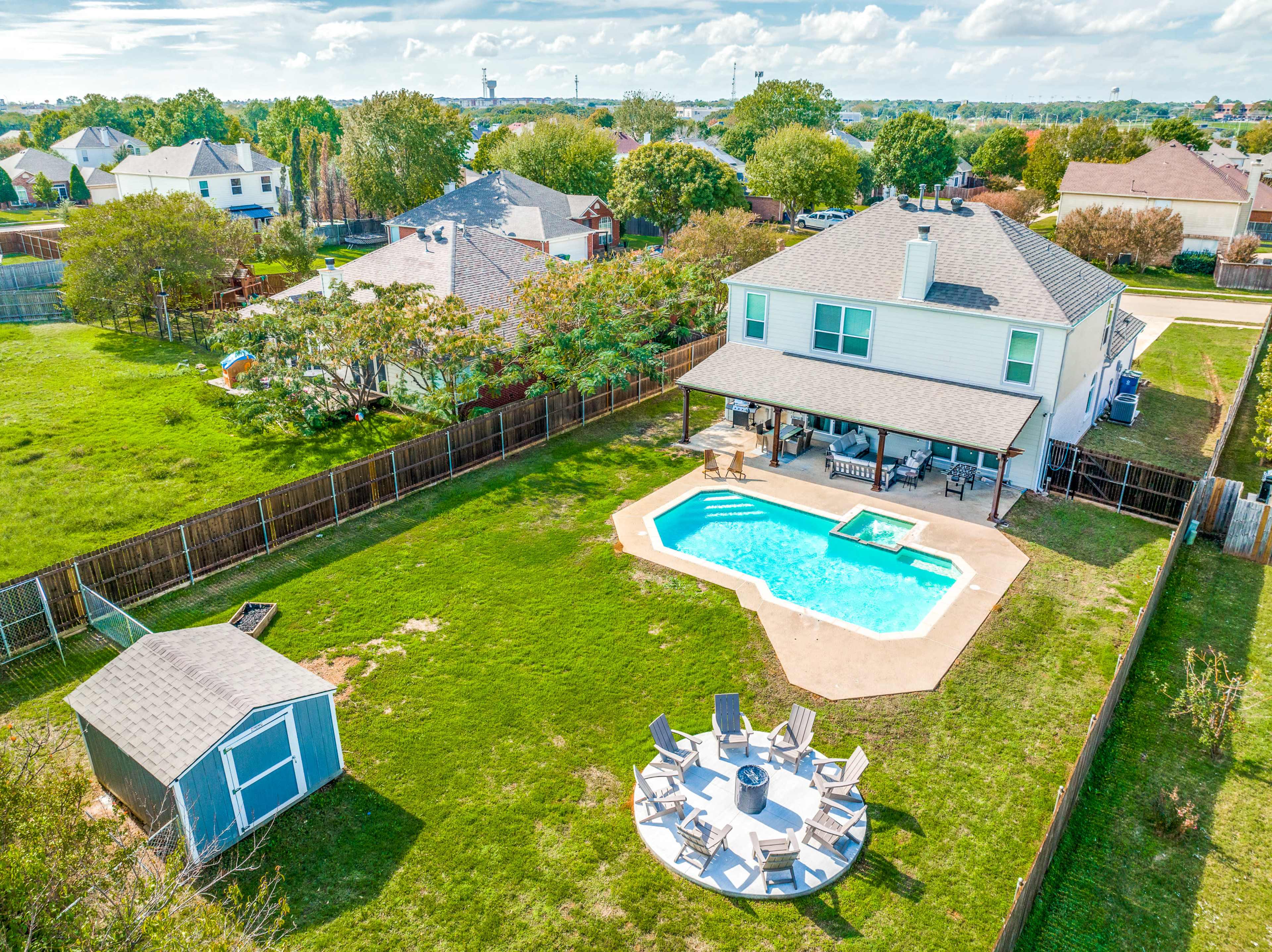 A backyard with a swimming pool, seating area, and a small shed, surrounded by a wooden fence and residential buildings in the background.