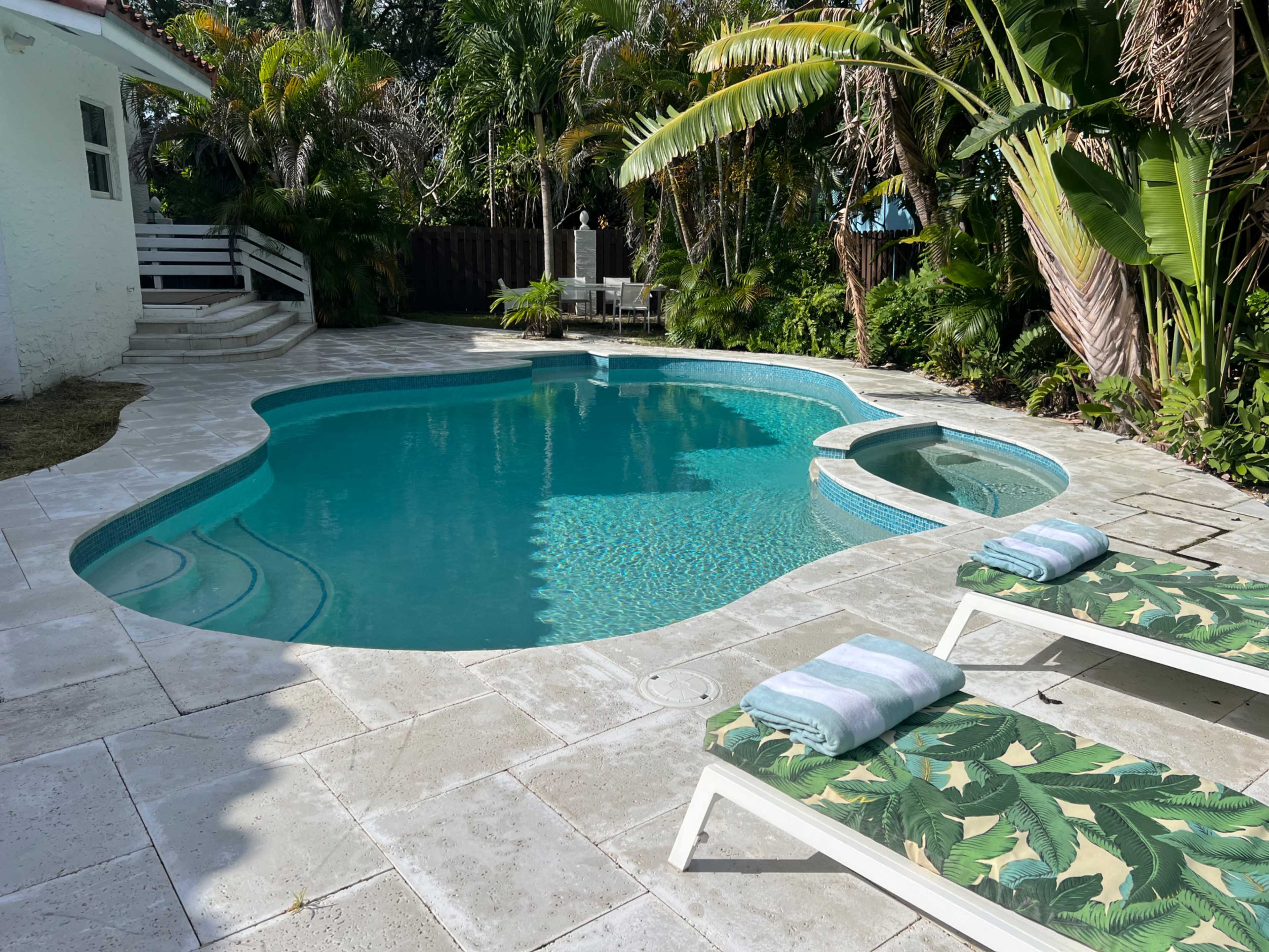 A backyard features a swimming pool surrounded by tropical plants and two lounge chairs with green and white leaf-patterned cushions.