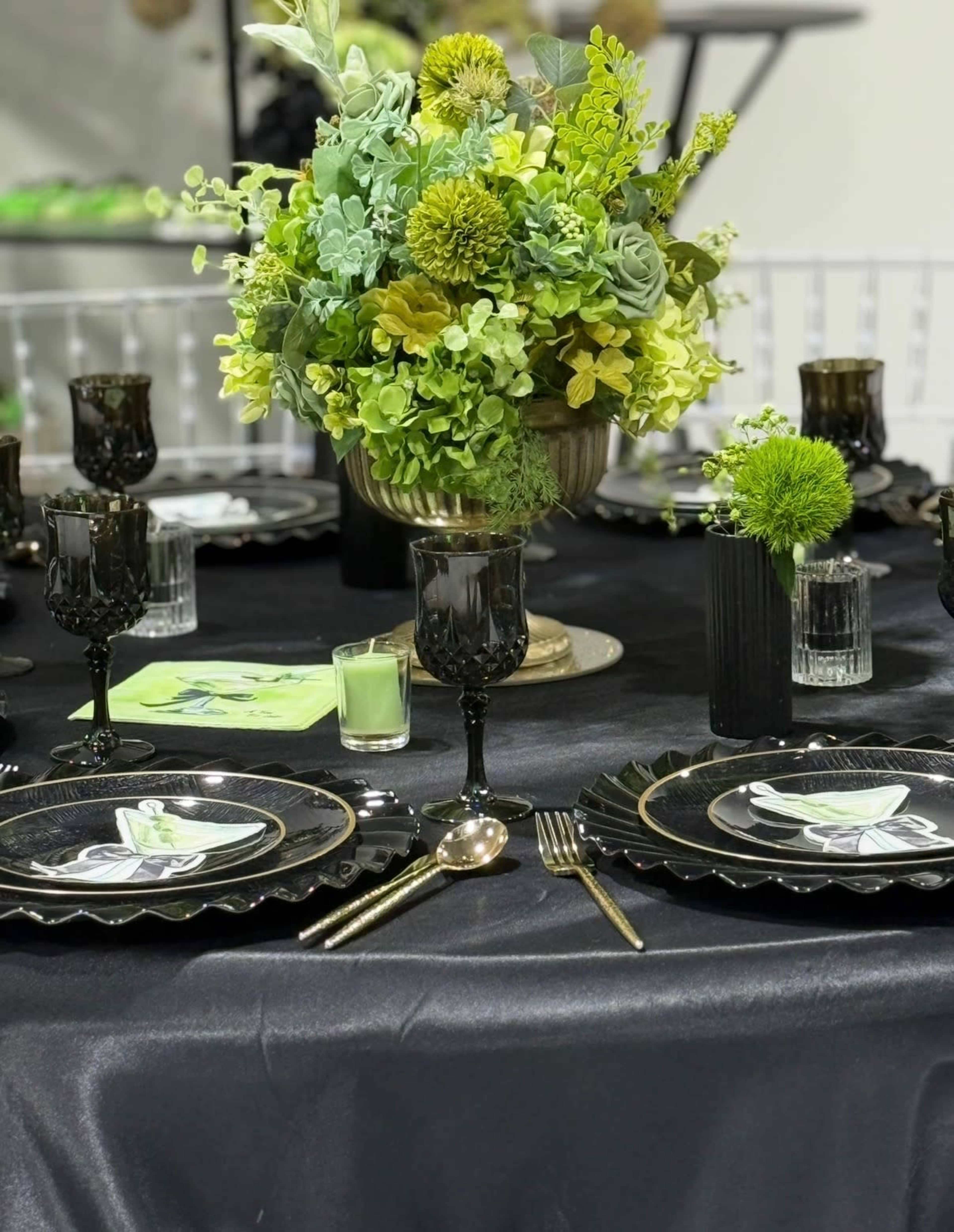 The image features a dining table set with black tableware, gold utensils, and a centerpiece of green flowers and foliage.